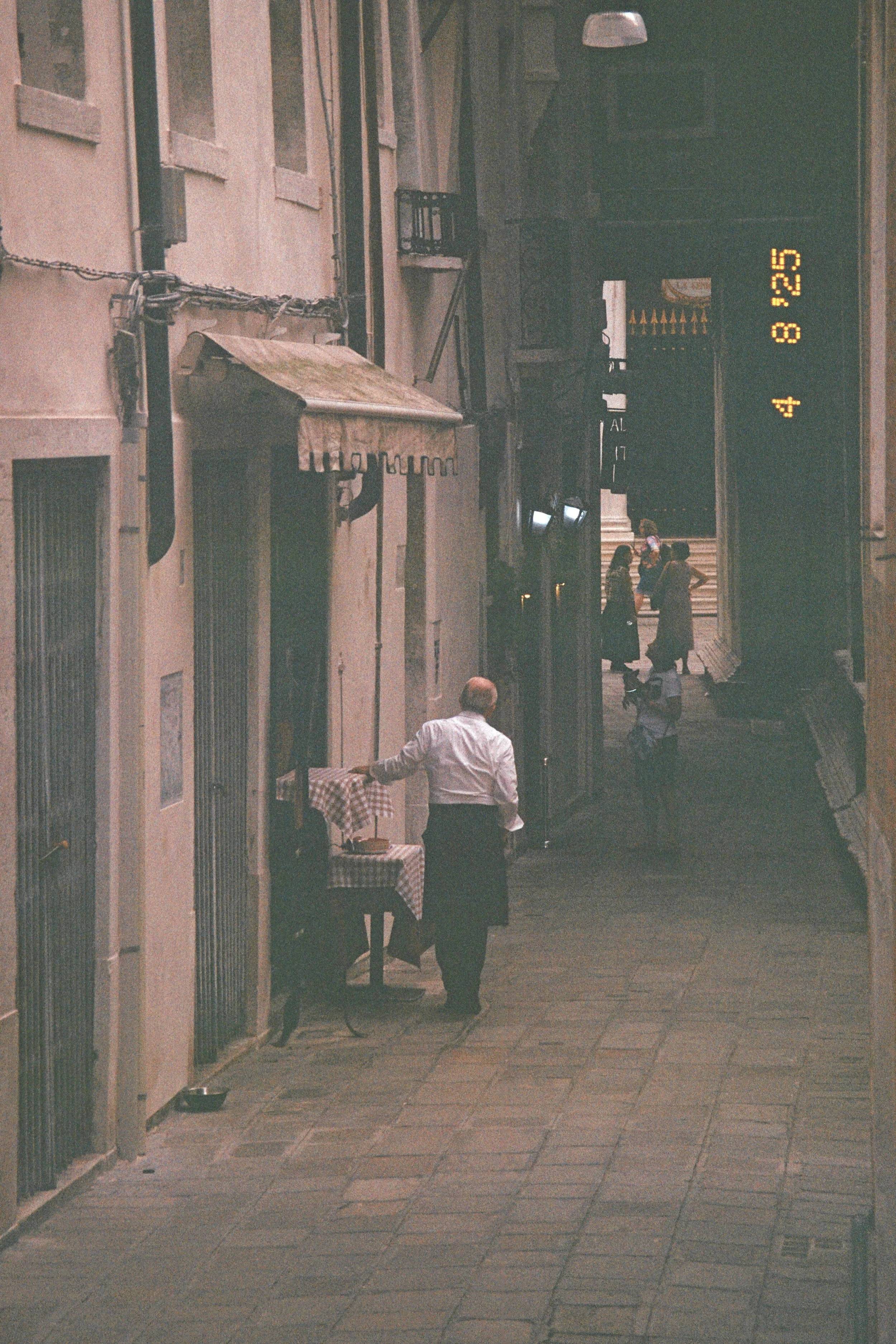 A man in a white shirt stands on a quiet, narrow street with outdoor tables outside small shops, and a few pedestrians walking in the distance, with a large digital clock displaying the time and date.