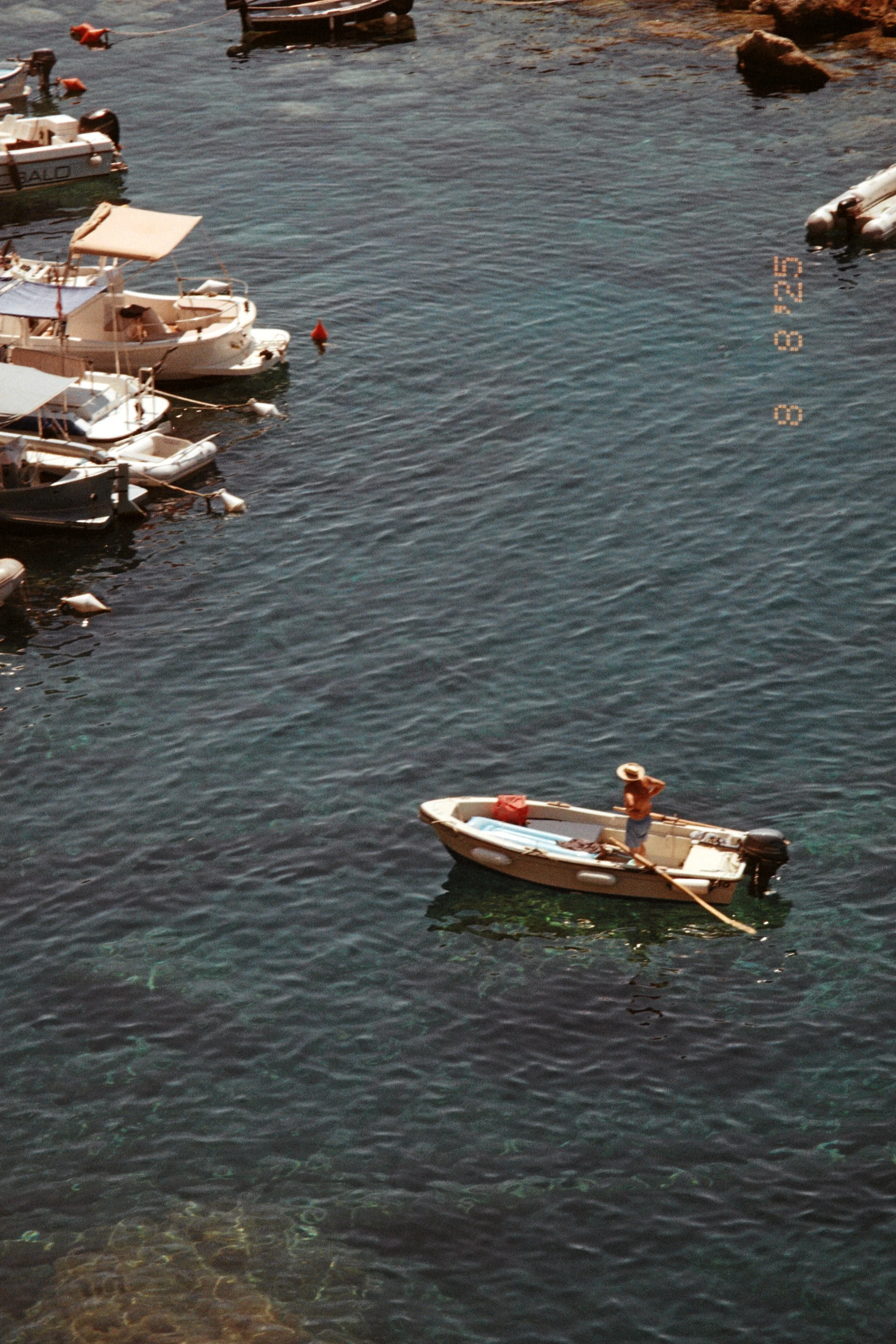 A person standing on a small boat in a marina, surrounded by larger boats moored to the dock.