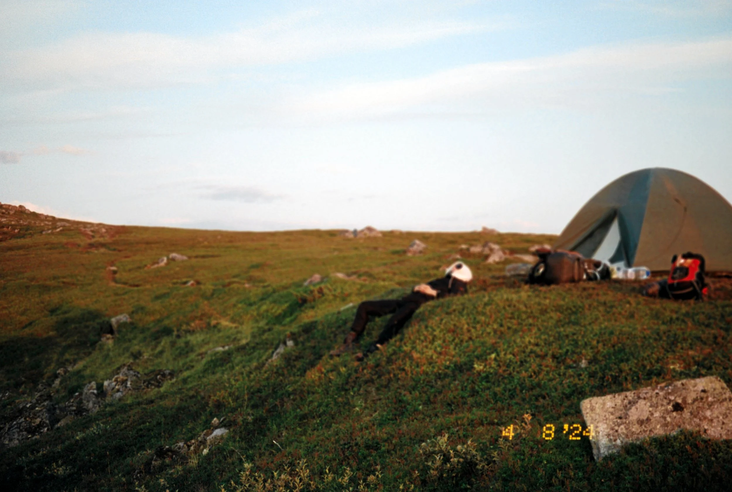 A person lying on the grass near a tent on a hillside with rocks, under a partly cloudy sky.