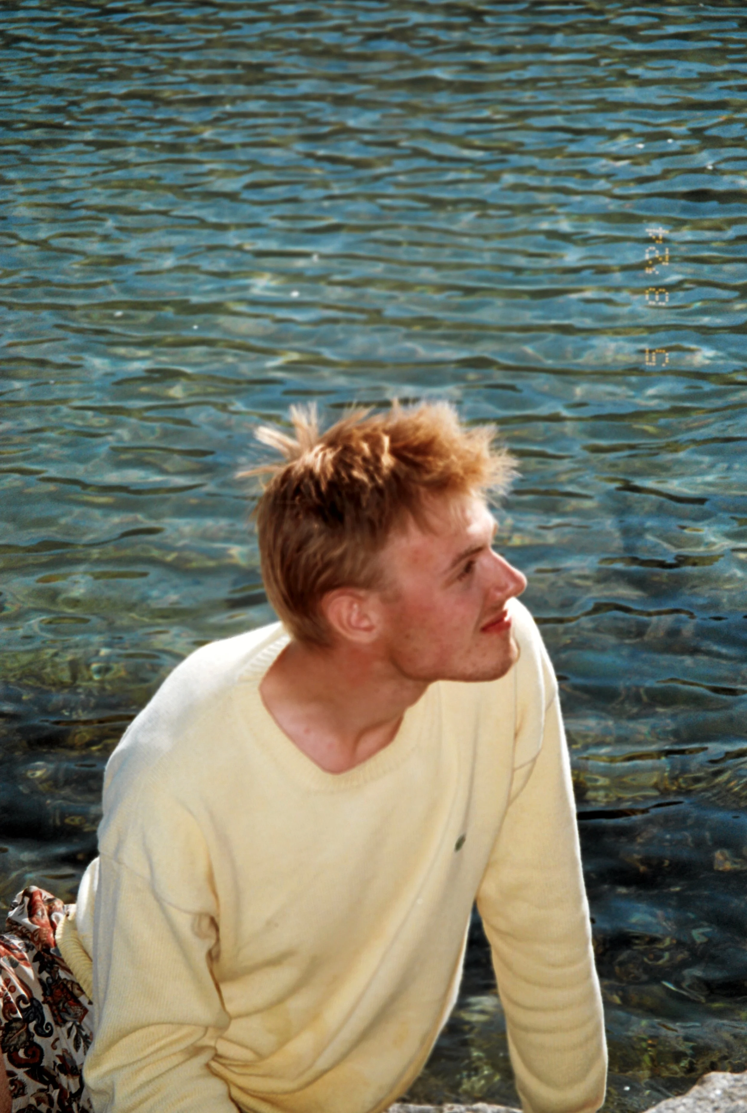 A young man with short, tousled light brown hair, wearing a cream-colored sweater, sitting by the edge of a body of water, looking to his right with a thoughtful expression.