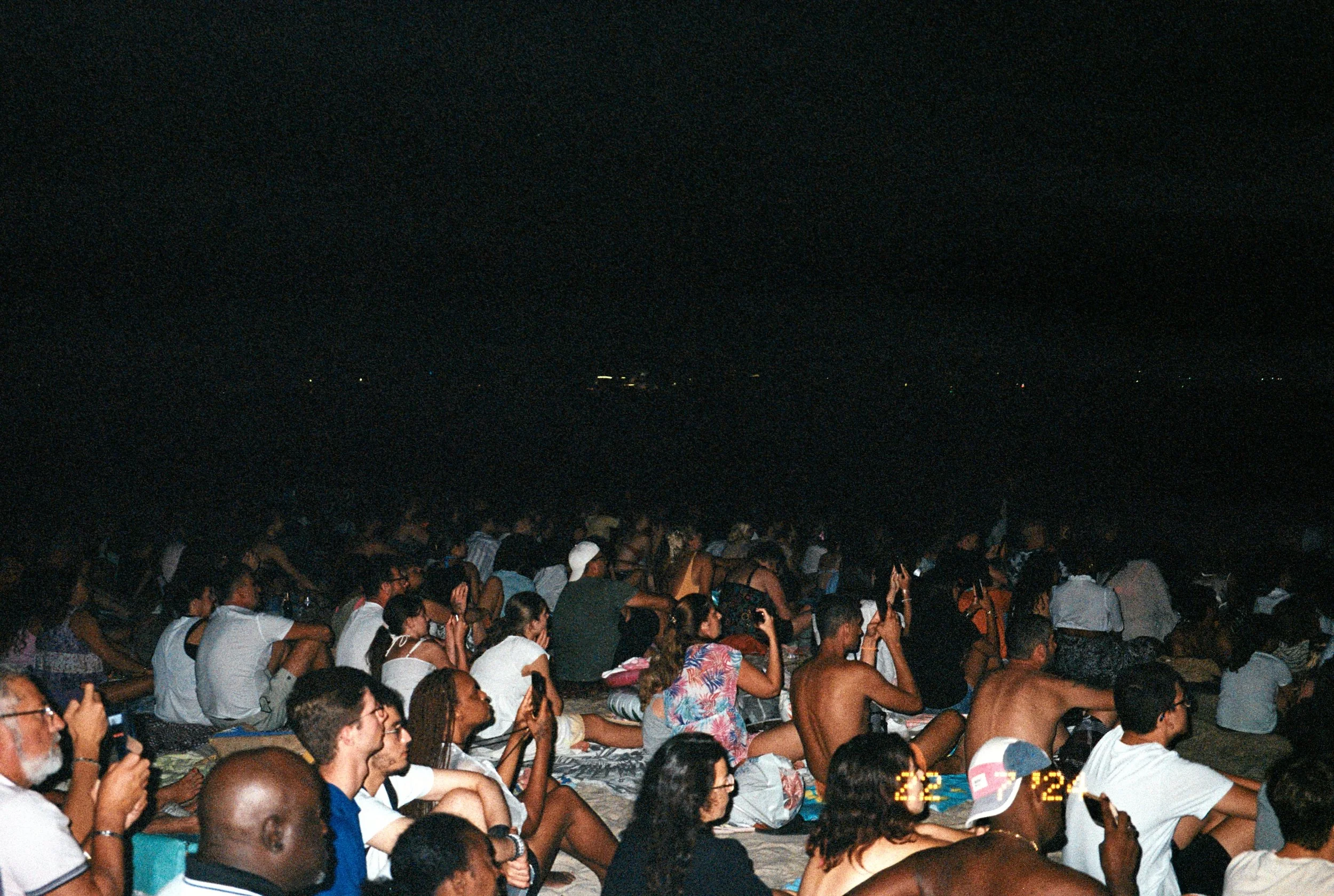 A large group of people sitting on the beach at night, facing the water, some taking photos or videos, with darkness and water in the background.