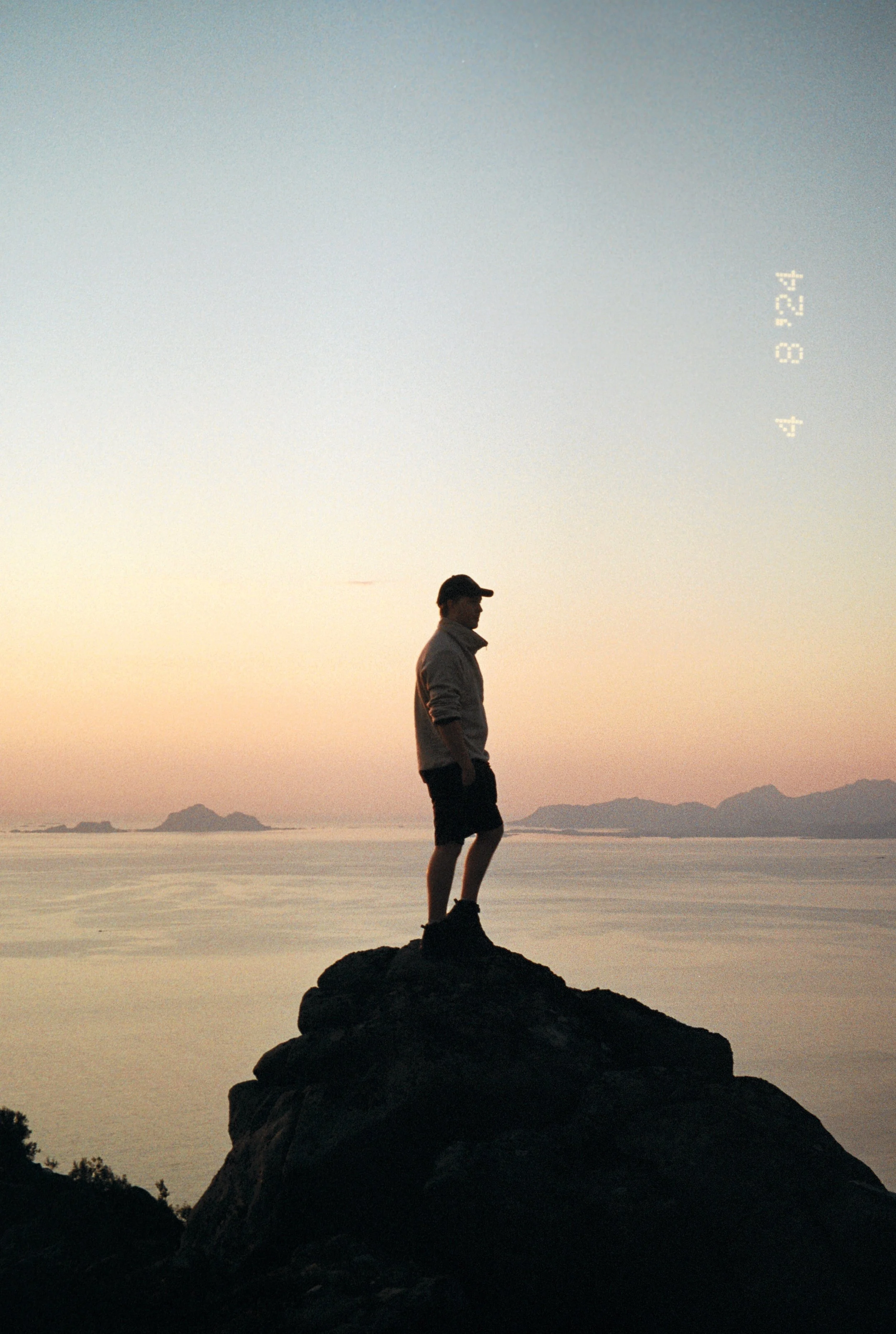 Silhouette of a person standing on a rocky outcrop at sunset, overlooking a calm body of water with distant mountains on the horizon.