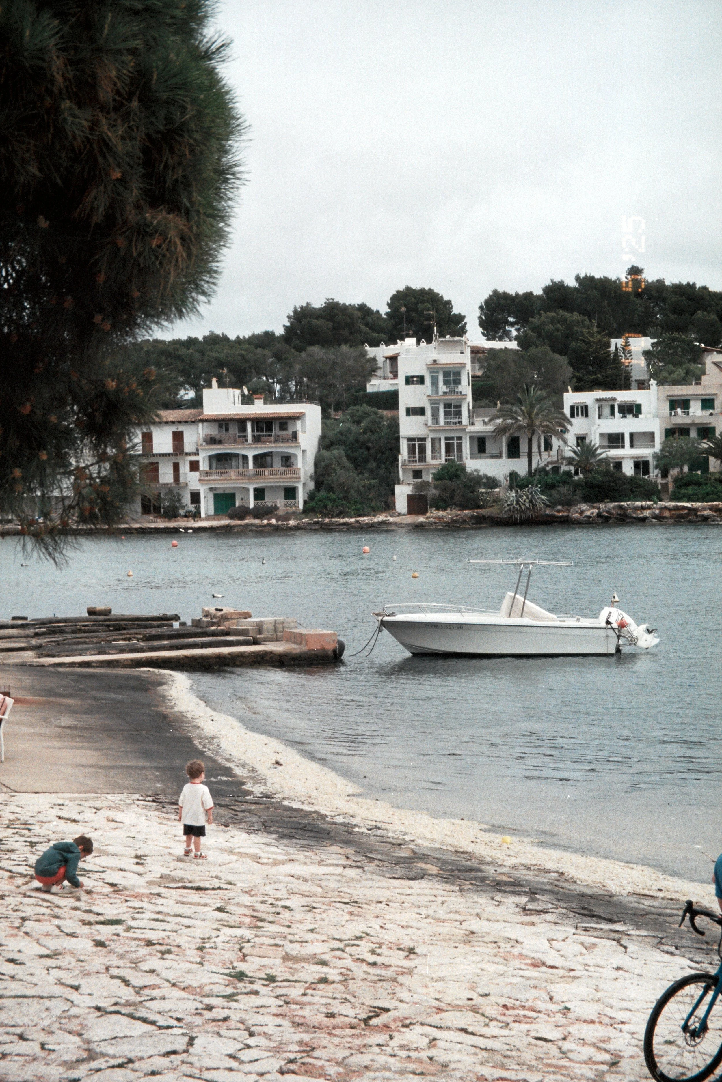 A peaceful lakeside scene with two small children playing on the stone shore, a boat moored in the water, and white residential buildings on a hill in the background.