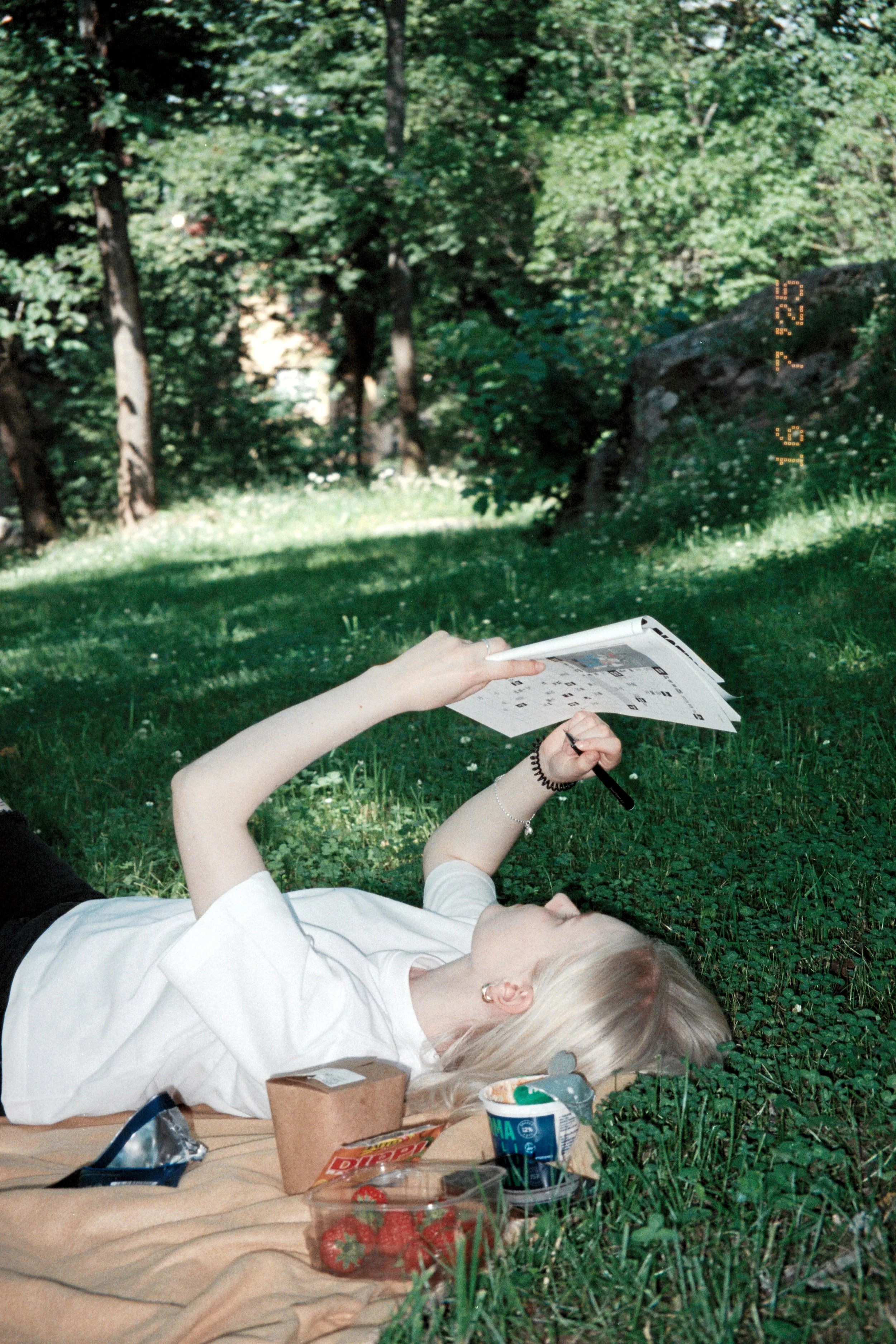 A person with platinum blonde hair, wearing a white shirt, is lying on the grass in a wooded area, reading a magazine or catalog. They are surrounded by various food and drink containers, including a cup, a box, and a plastic container, on a blanket.