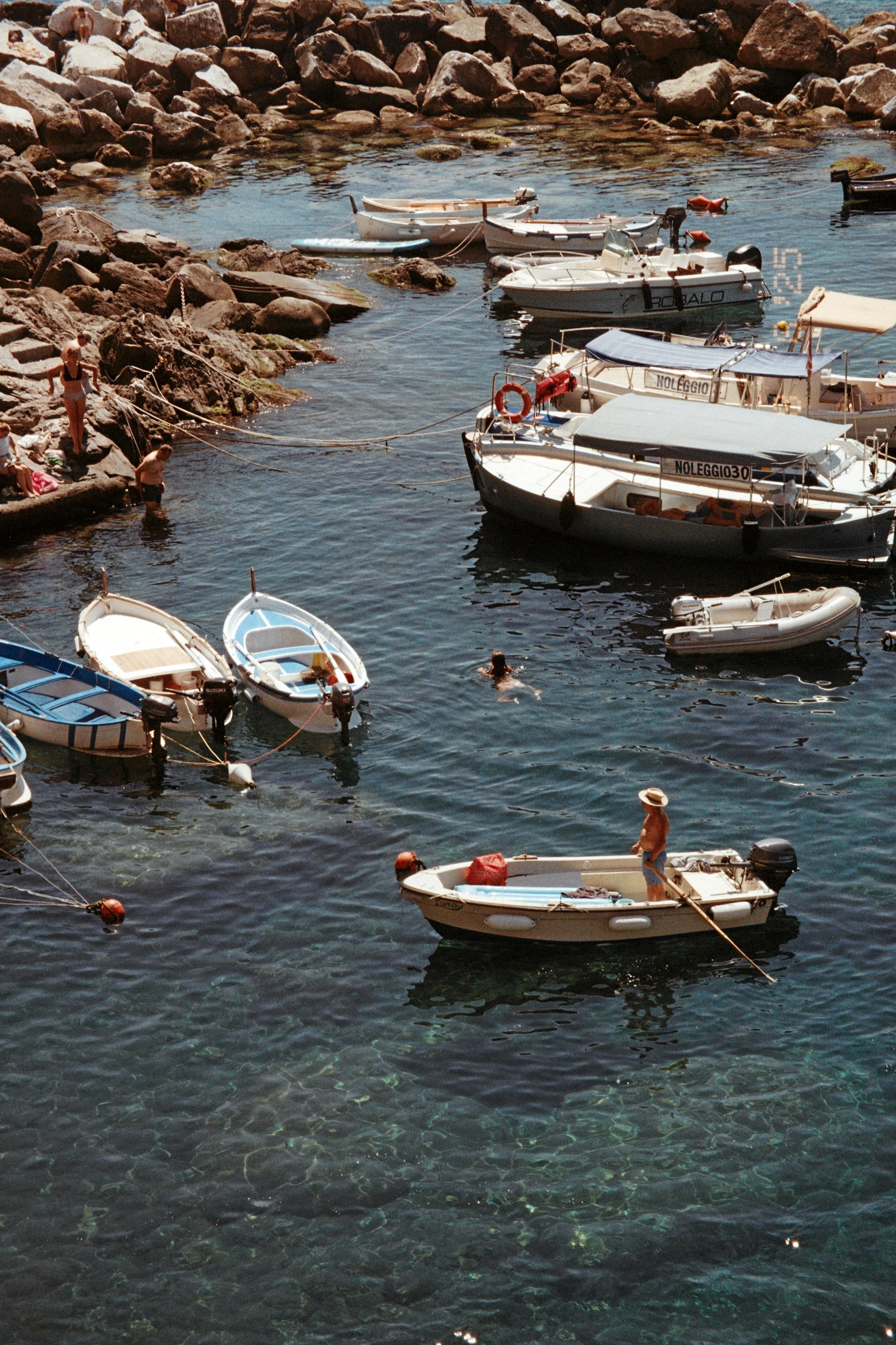People swimming and relaxing in a small rocky harbor with several moored boats, including small motorboats and sailboats, on a sunny day.