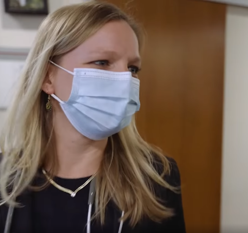 A woman wearing a light blue face mask, black shirt, gold jewelry, and with blonde hair, in an indoor setting with wooden walls.