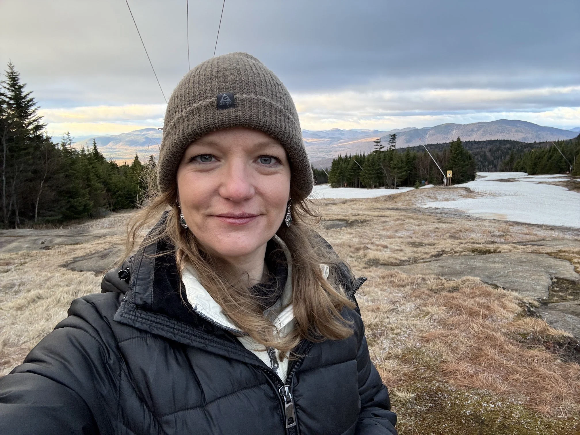 A woman with blonde hair and blue eyes wearing a brown knit cap, black jacket, and earrings taking a selfie outdoors in a mountainous landscape with trees, patches of snow, and cloudy sky.