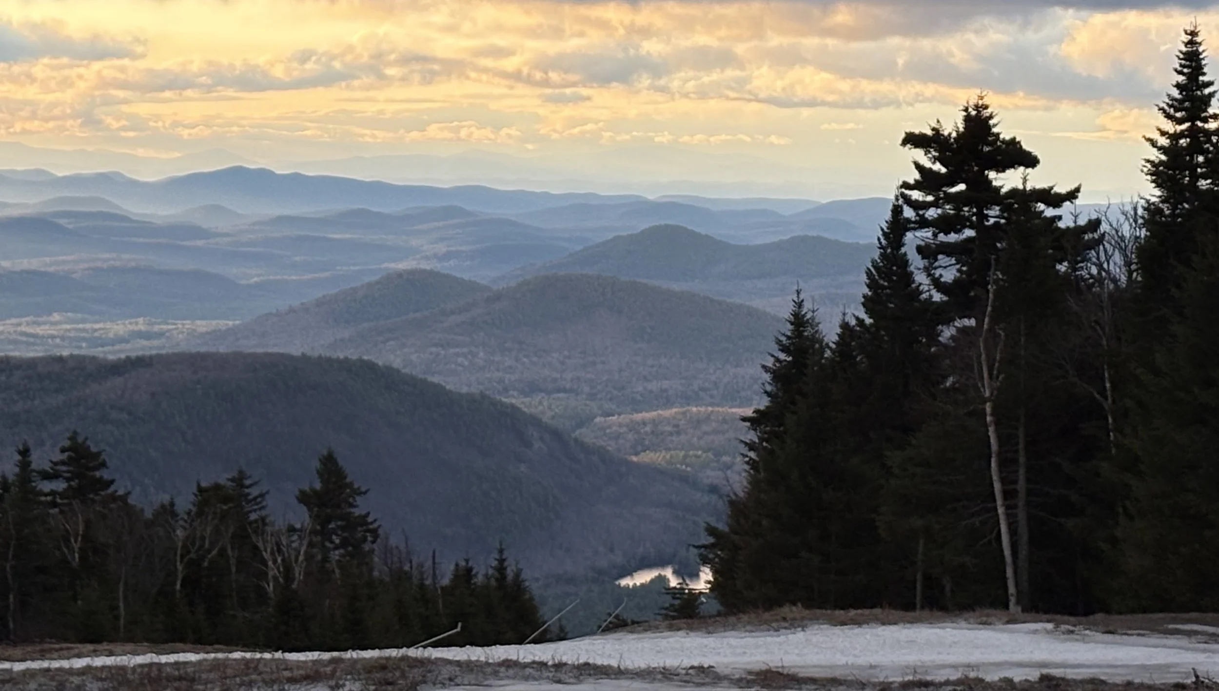 A scenic view of rolling mountains with layered blue hues, pine trees on the right, and a partly cloudy sky at sunset or sunrise.