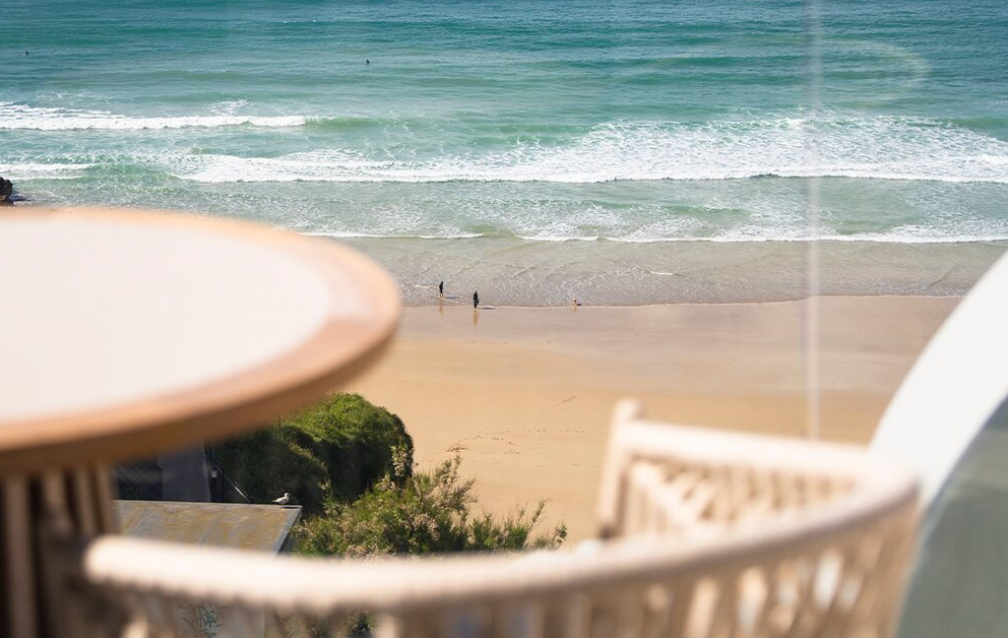 View of a beach with ocean waves, sandy shore, and two people walking along the water's edge seen from a balcony with blurred chairs and a table in the foreground.