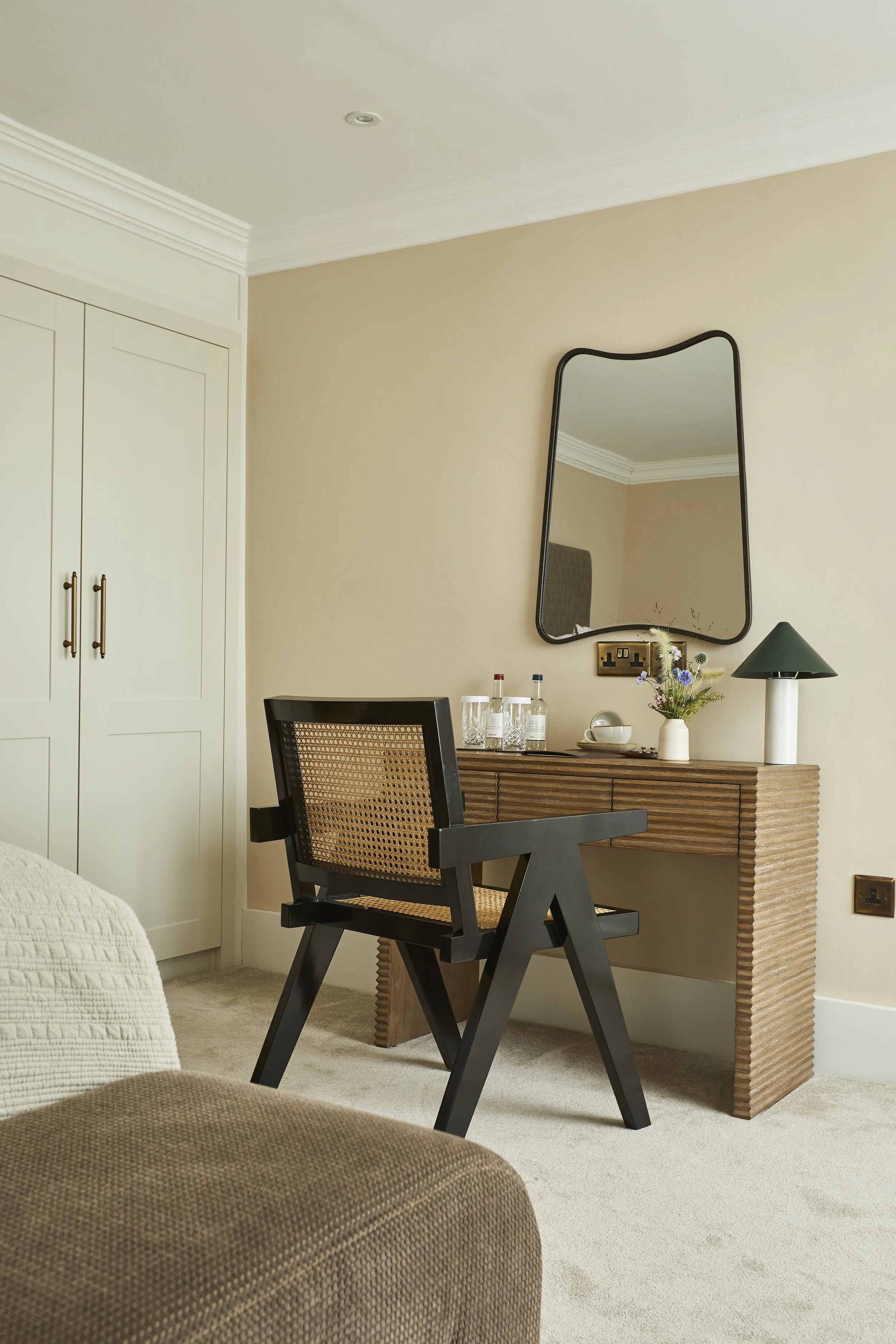 A living room with a beige wall, a dark wooden chair with rattan backrest, a wooden vanity table with decorative textural details, a mirror, a green lampshade, and a small vase with flowers.