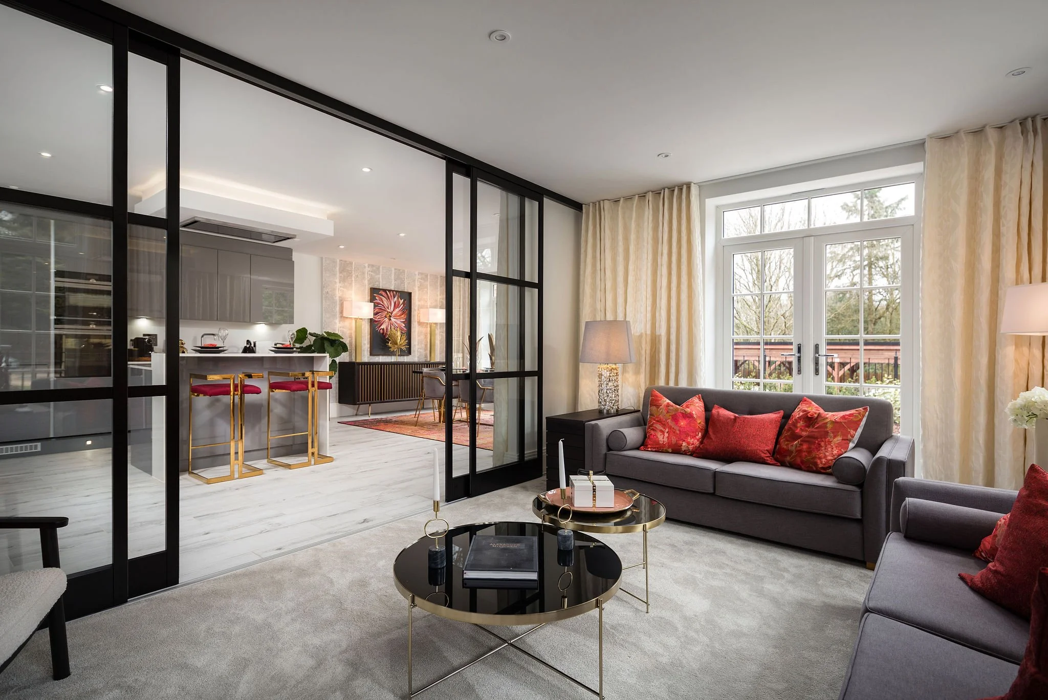 Living room with sofas, pillows, coffee table, and floor lamps, separated from kitchen and dining area by glass partition, with large windows and curtains.
