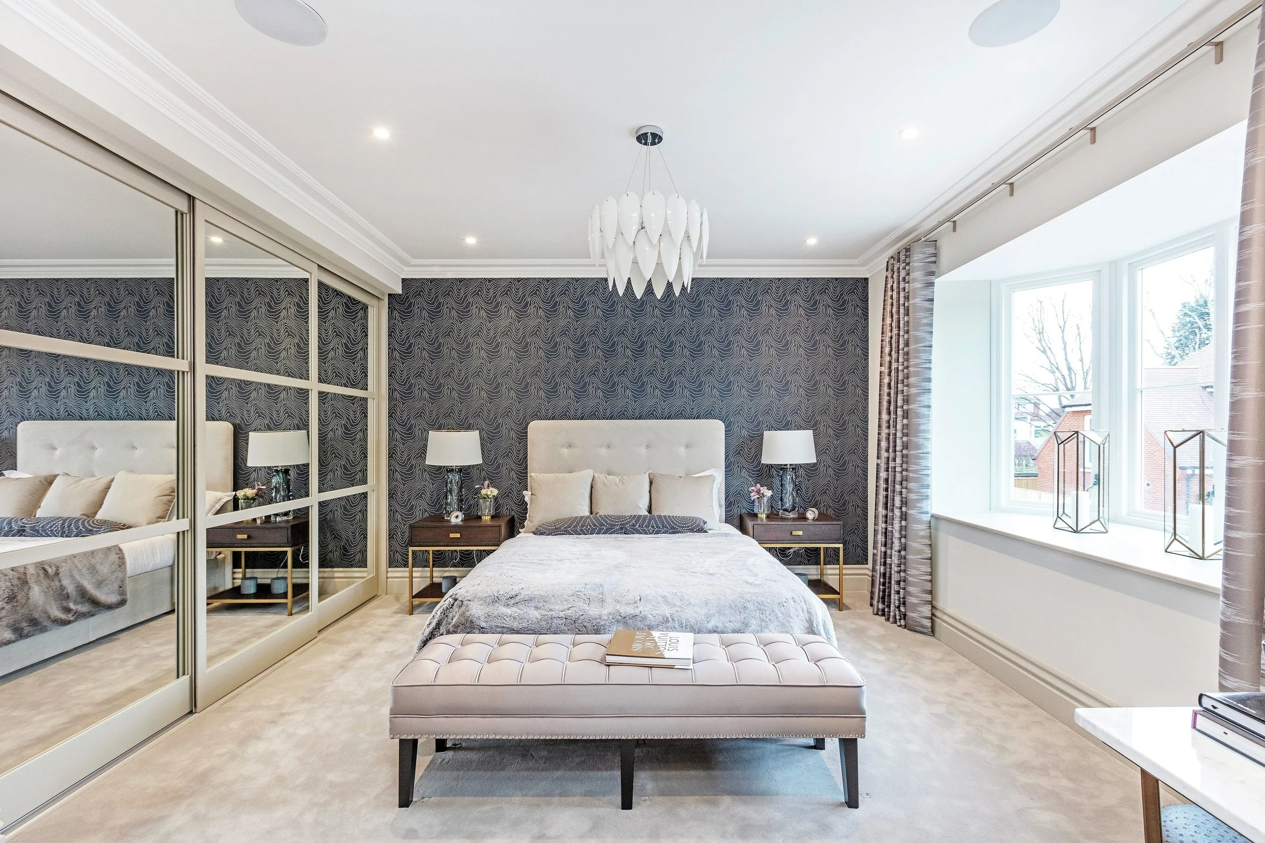 A modern bedroom features a large bed with white and beige pillows, dark wood nightstands with white lamps, a wall with dark patterned wallpaper, a mirrored closet, and a window with curtains overlooking snowy houses.