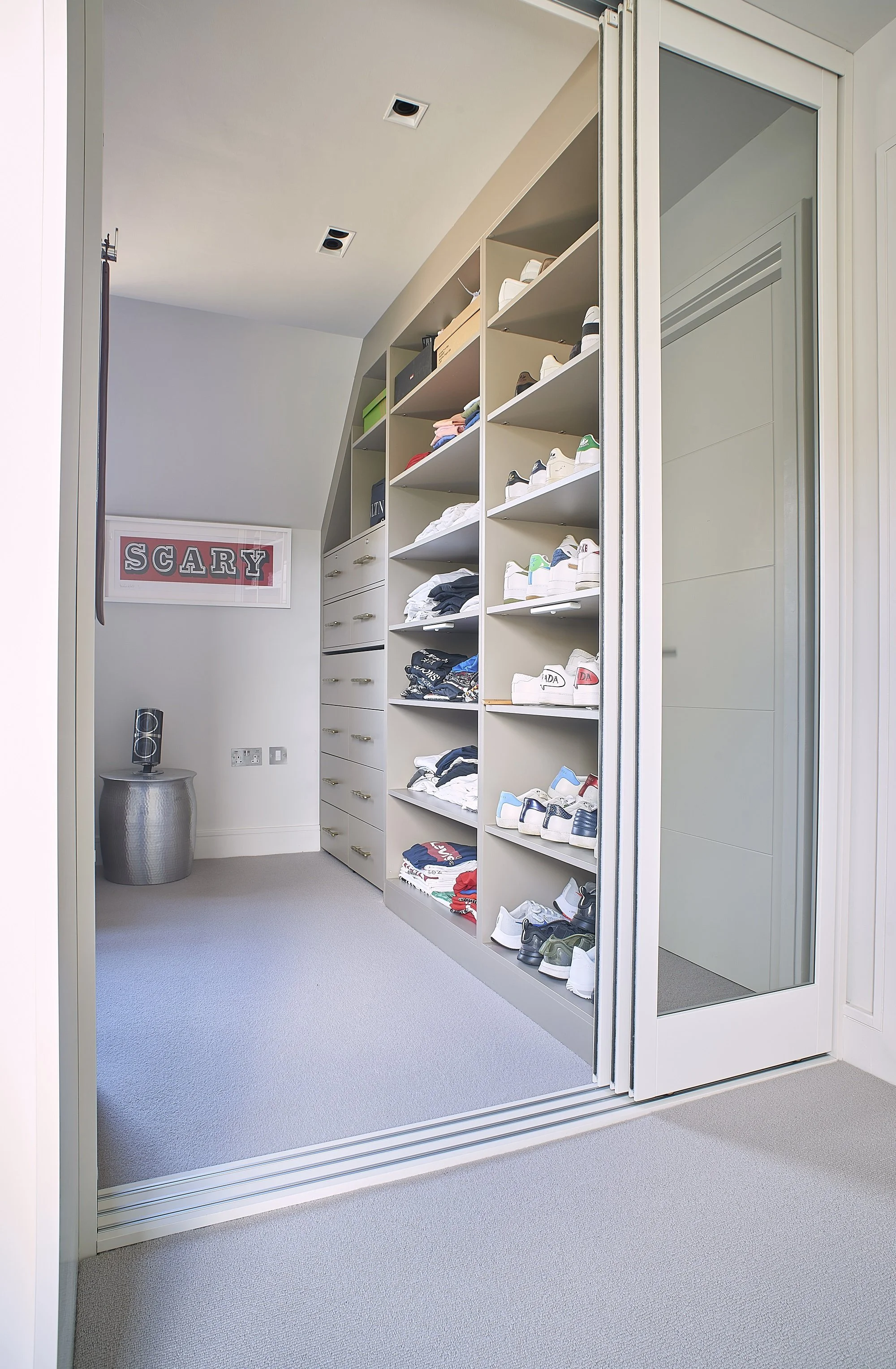 A walk-in closet with open shelving filled with neatly folded clothes and shoes, a mirrored sliding door, a framed 'SCARY' poster on the wall, and a small metallic table with a stereo on top.