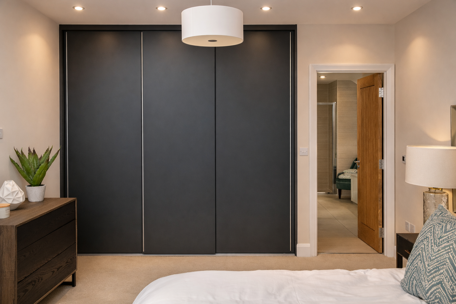 A bedroom with a large black closet, an open wooden door leading to a bathroom, a wooden dresser with a potted plant, a nightstand with a lamp, and a bed with patterned pillows.