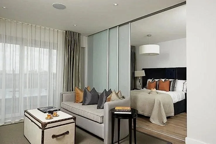 Modern hotel room with a seating area, glass wall divider, and a bedroom with a bed, nightstand, and hanging ceiling light.