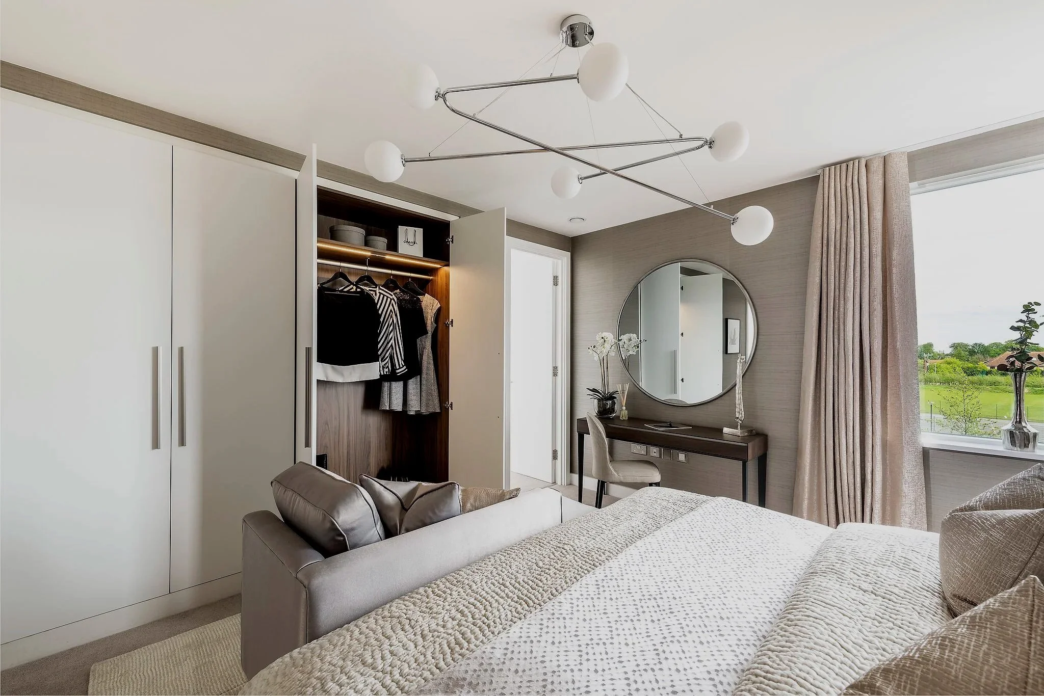 Modern bedroom with a bed, open closet with clothes, small desk with mirror, and window with curtains showing a green outdoor scene.