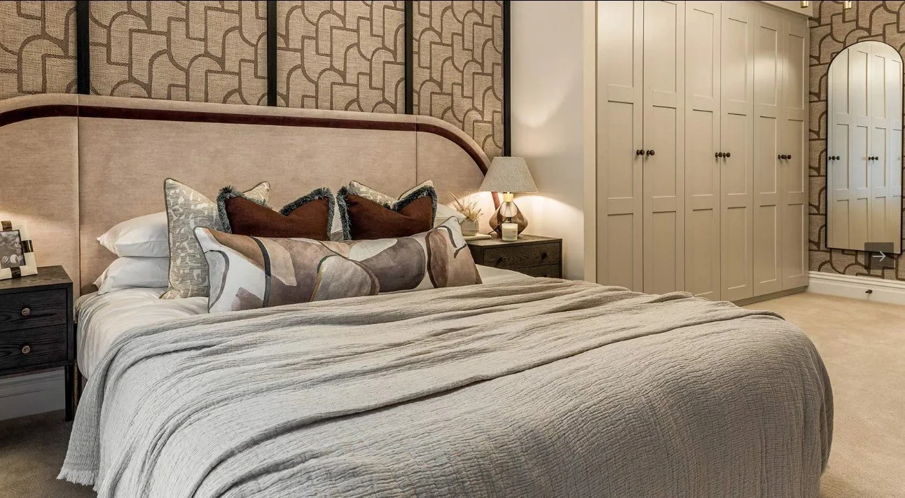 A cozy bedroom with a large bed, three decorative pillows, two nightstands with lamps, a patterned accent wall, and a large white closet with multiple doors.