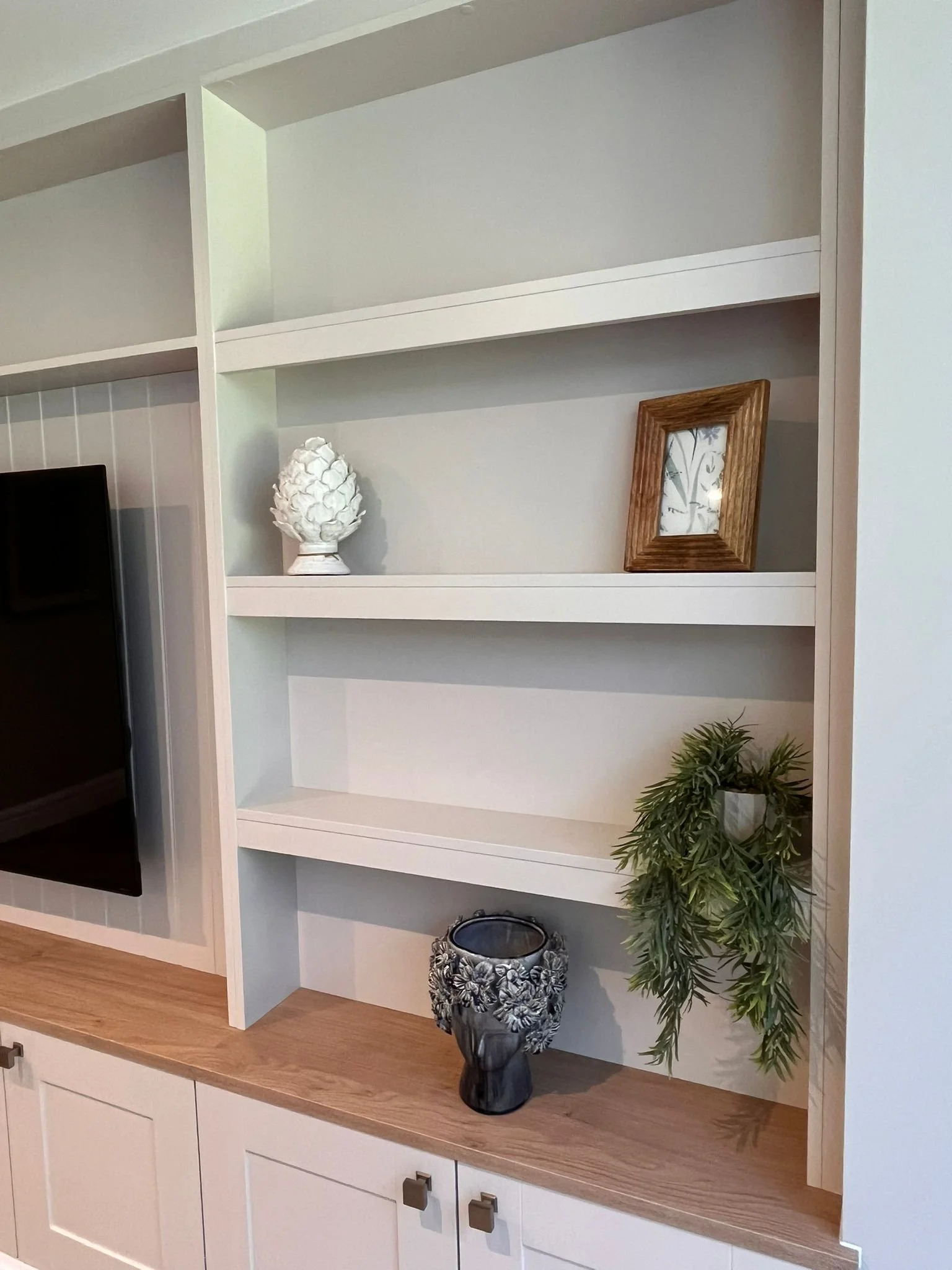 A white built-in shelf with three decorative items: a white artichoke sculpture, a wooden picture frame, and a hanging green plant. The shelf is part of a larger white cabinet with closed storage below.