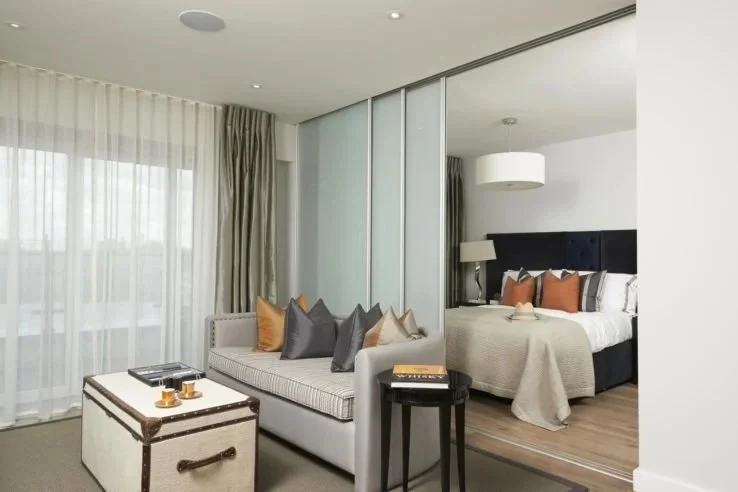 Modern hotel suite with a sitting area and a bedroom separated by a glass wall and sliding doors. The sitting area has a white sofa with multiple colorful pillows, a small black side table, and a white trunk used as a coffee table. The bedroom features a large bed with white linens, a navy blue headboard, a nightstand with a lamp, and decorative pillows. Large windows with sheer curtains allow natural light into the room.