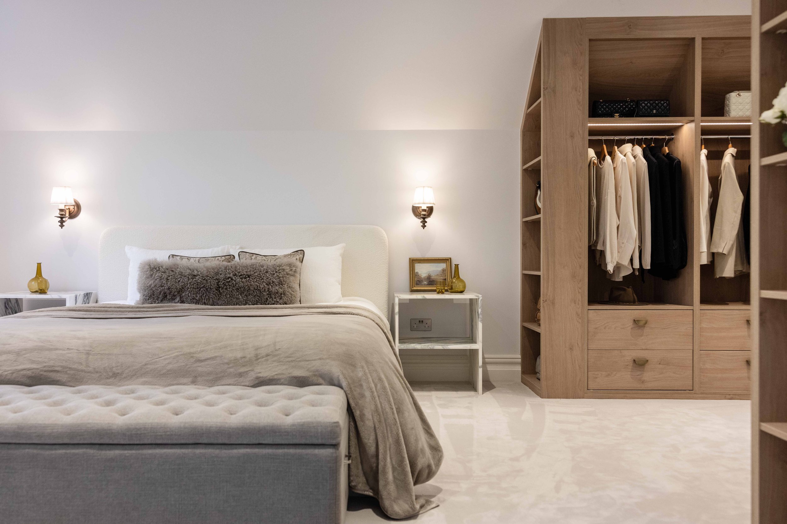 A bedroom with a white bed and gray pillows, a small white nightstand with a lamp, and an open wooden closet with clothes hanging inside.