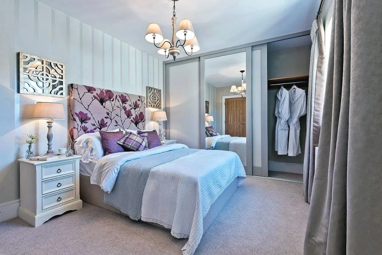 A cozy bedroom with a large bed, floral headboard, bedside tables with lamps, mirrored closet doors, and gray curtains, decorated in soft pastel colors.