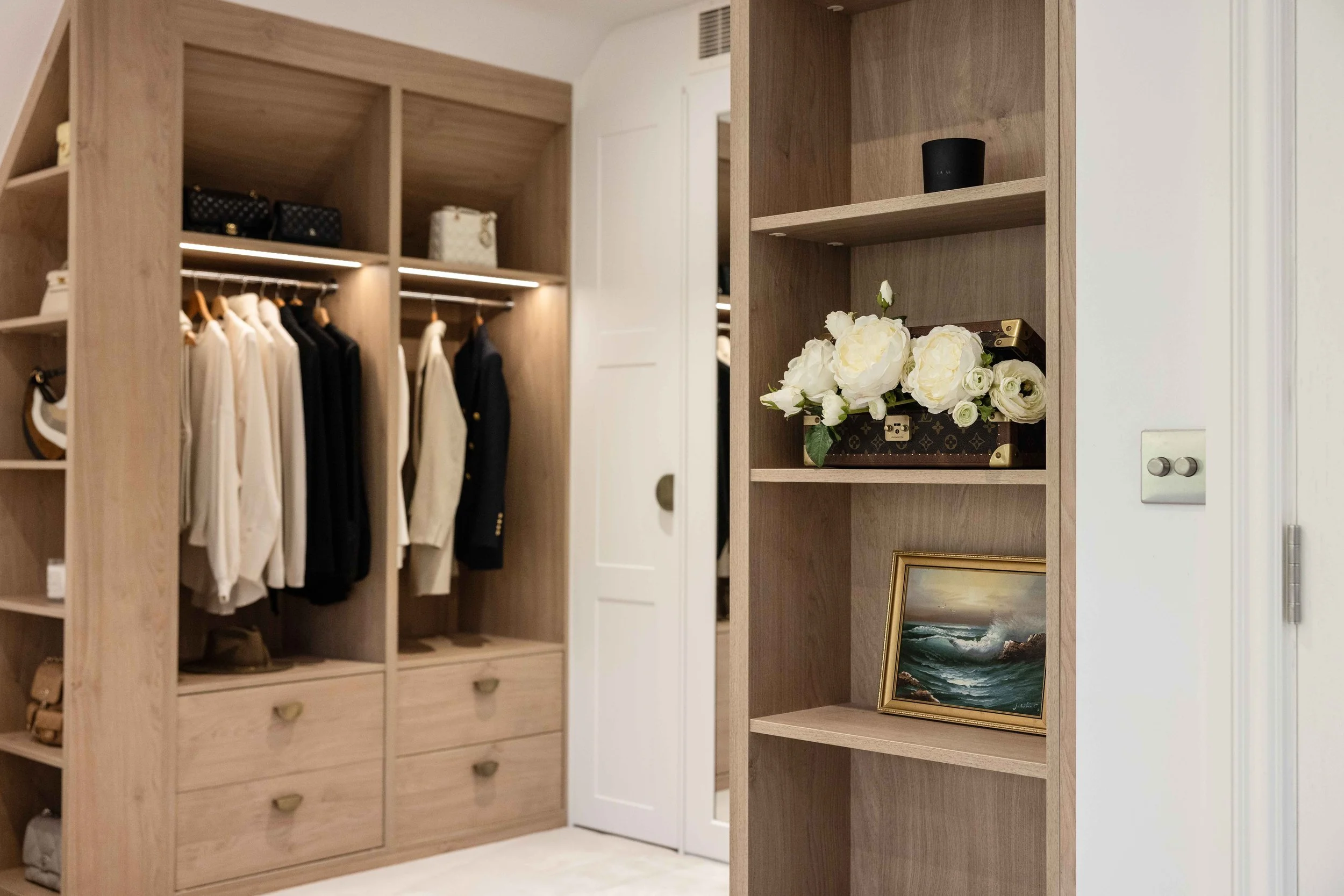 A walk-in closet with wooden shelves and cabinets, displaying clothes on hangers, handbags, and decorative items including flowers and a framed painting.