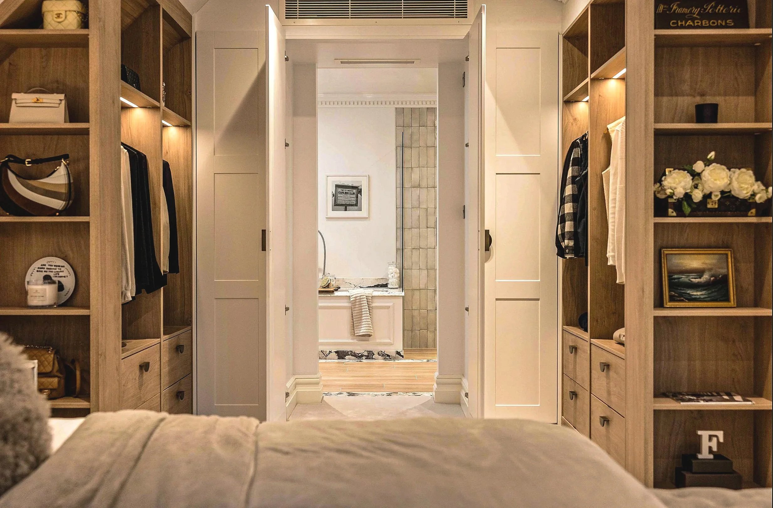 A view from a bed looking towards an open doorway leading to a bathroom, with built-in wooden shelving on each side filled with decorative items, clothing, and books.