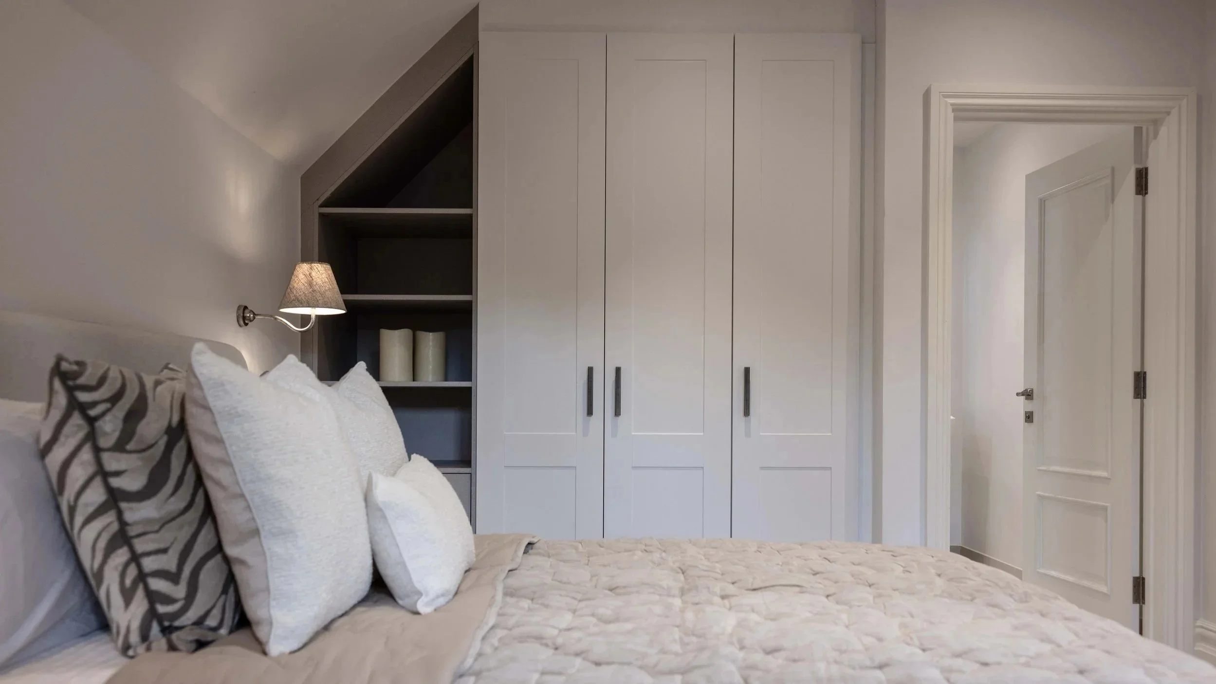 A bedroom with a bed featuring multiple white and patterned pillows, a wall-mounted lamp, a built-in wardrobe, and a door leading to a hallway.