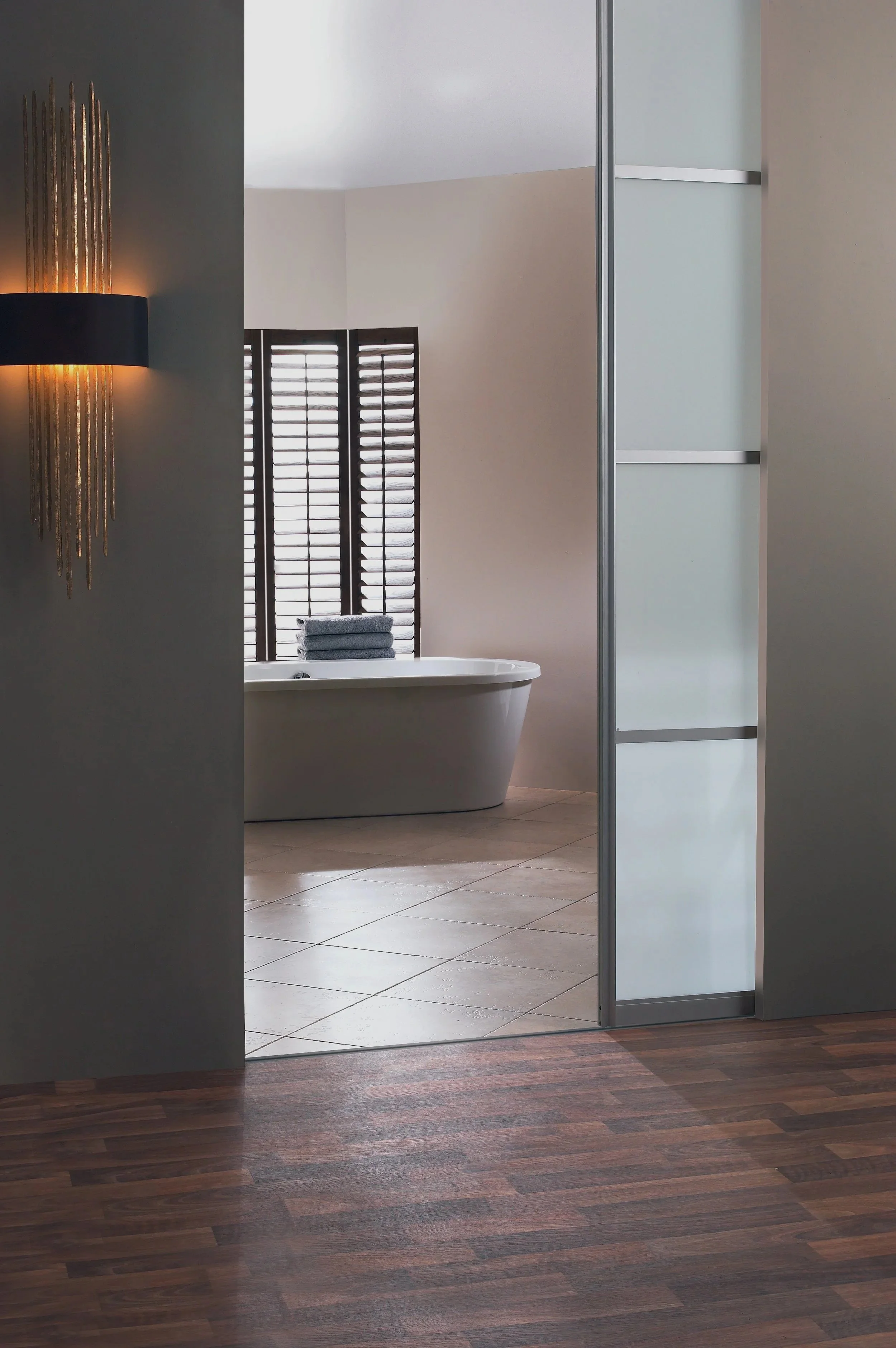 Modern bathroom with a bathtub, wooden blinds, towels, and a decorative wall lamp.