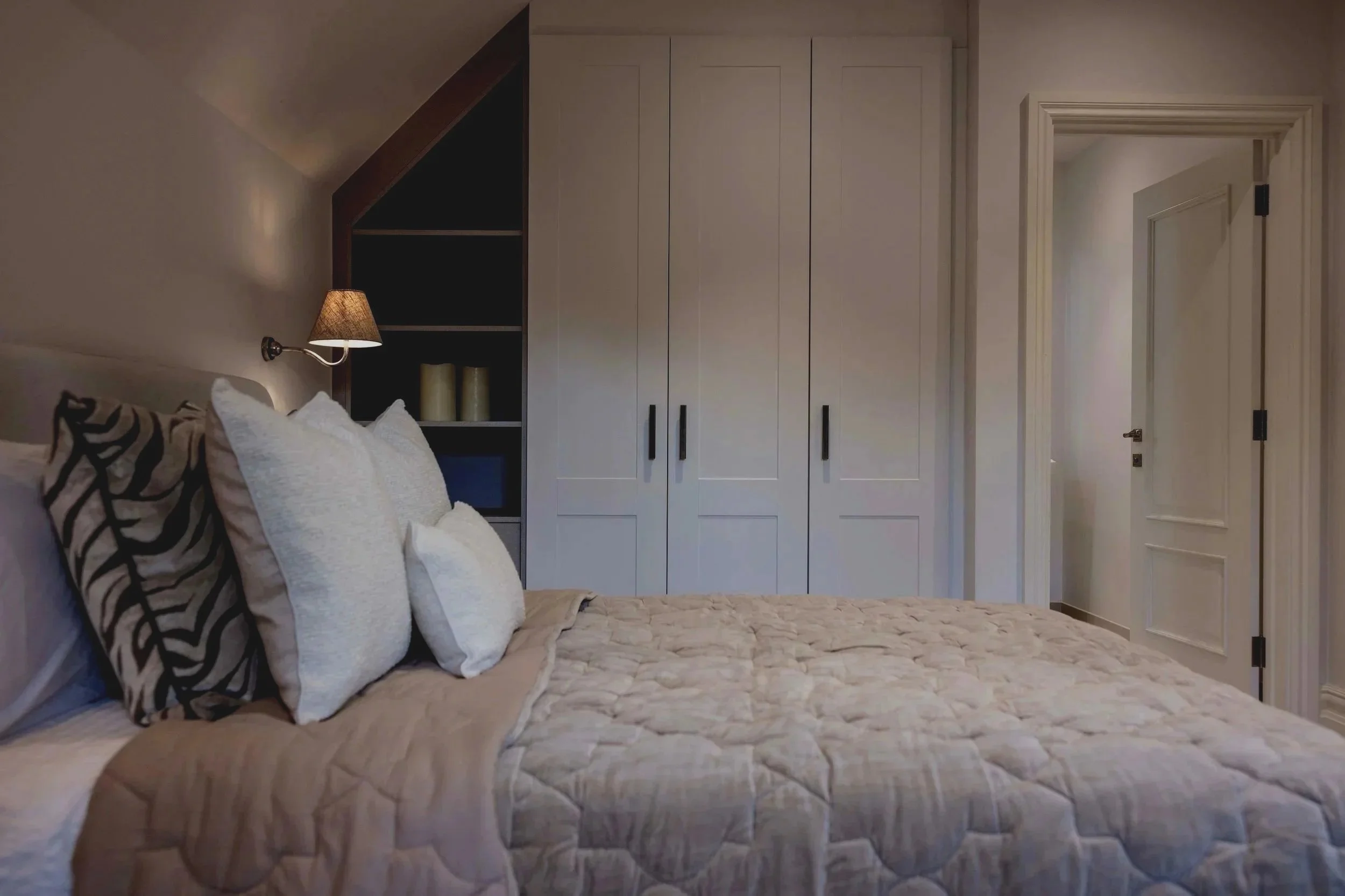 A bedroom with a bed, pillows, wall lamp, and a large closet with white doors. There is an open door leading to a hallway.
