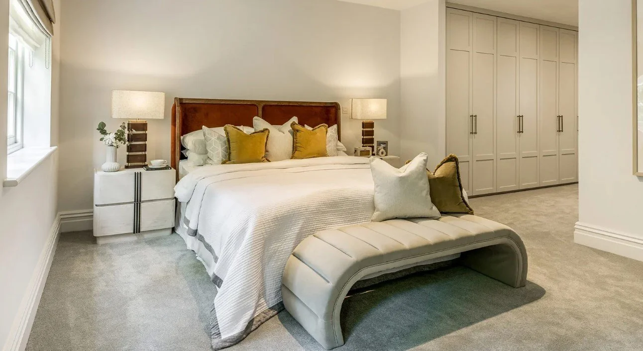 A cozy bedroom with a large bed, white bedding, and decorative pillows. There are two nightstands with lamps on each side of the bed, a window with blinds, a bench at the foot of the bed, and a large closet with white doors in the background.