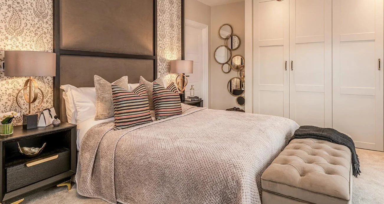 A bedroom with a large bed, striped pillows, beige bedding, lamps on bedside tables, decorative mirror on the wall, and a built-in closet