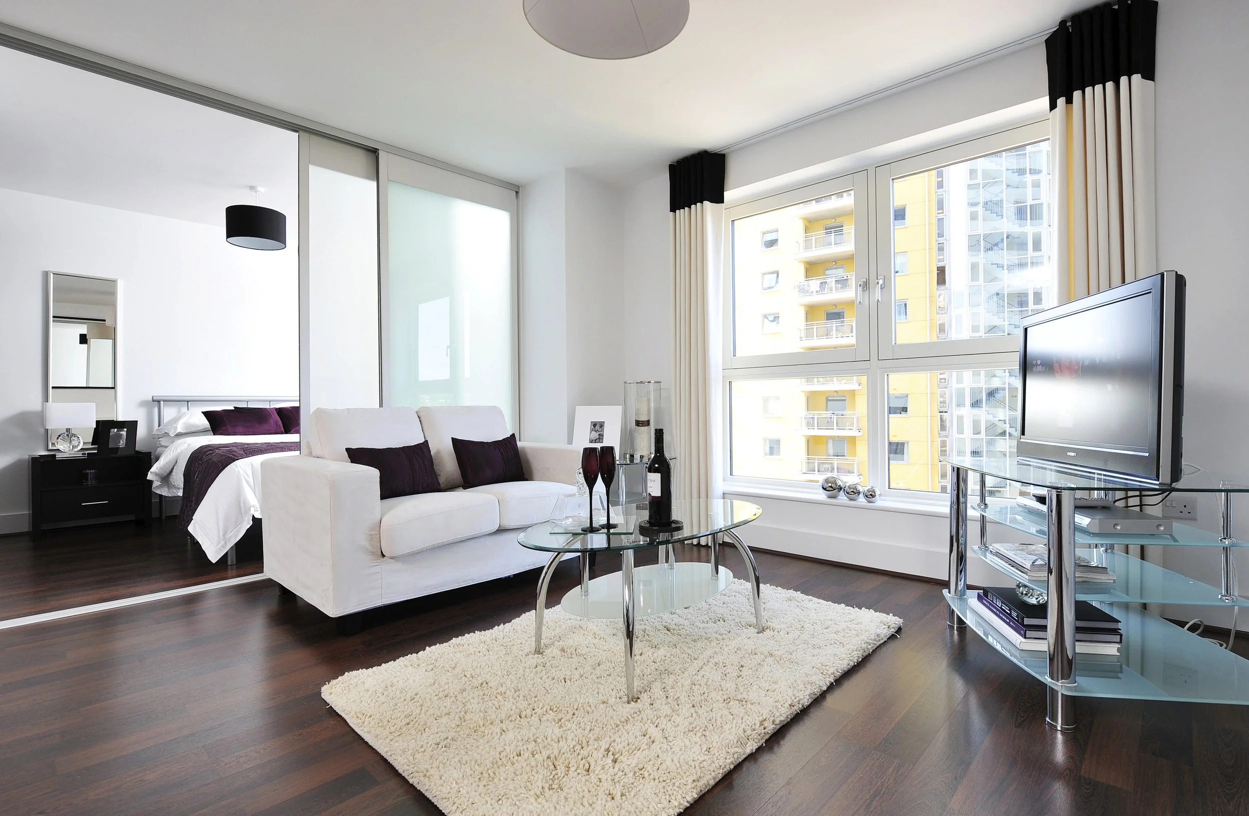 Modern living room with large windows, white sofa with purple pillows, glass coffee table with wine and glasses, area rug, and a TV stand with a flat screen TV.