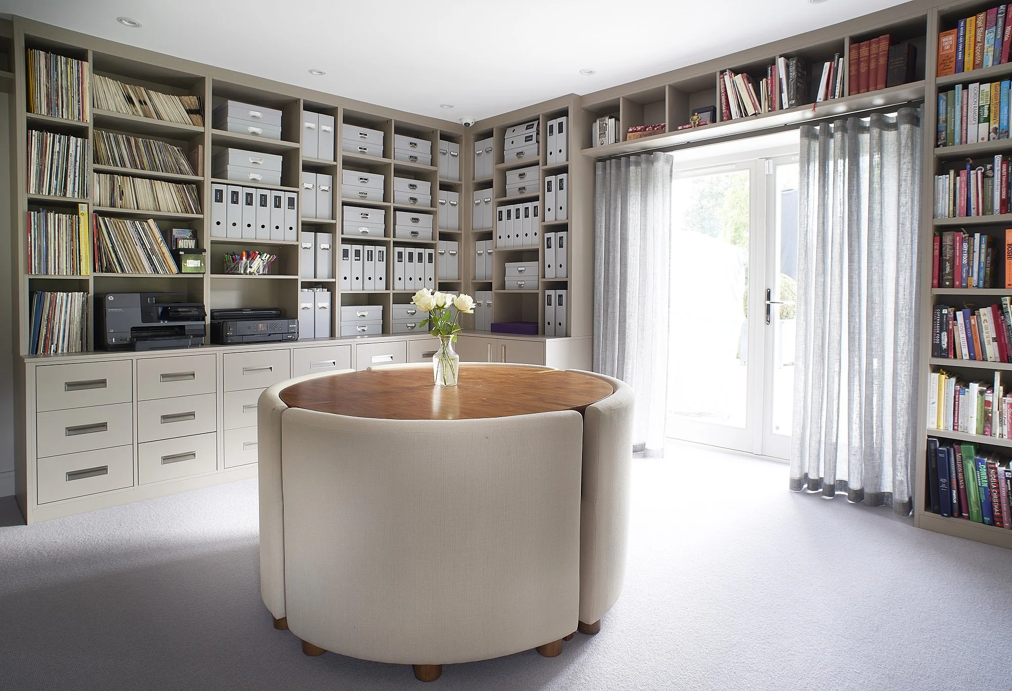 A room with built-in bookshelves filled with books, magazines, and file boxes, a round wooden table with a glass vase holding flowers, and a beige curved armchair. There are curtains covering glass doors leading outside, and a white carpet on the floor.