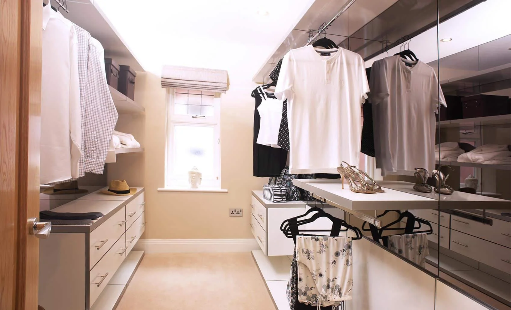 A walk-in closet with hanging clothes, shelves with folded garments, shoes on a surface, and a window with a beige shade.