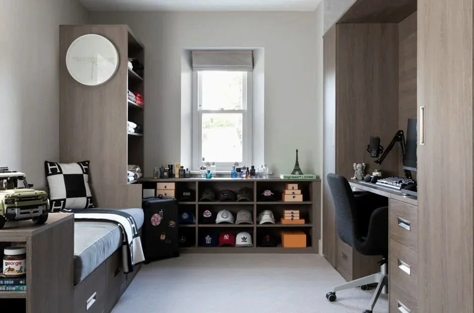 A bedroom with a single bed, desk, and storage units. The bed has a black and white striped blanket, pillows, and a toy car. The wardrobe and shelves are made of light wood. The window has blinds, and the shelves contain hats, candles, and decorative items like a small Eiffel Tower replica. The desk has a computer, monitor, and office chair.