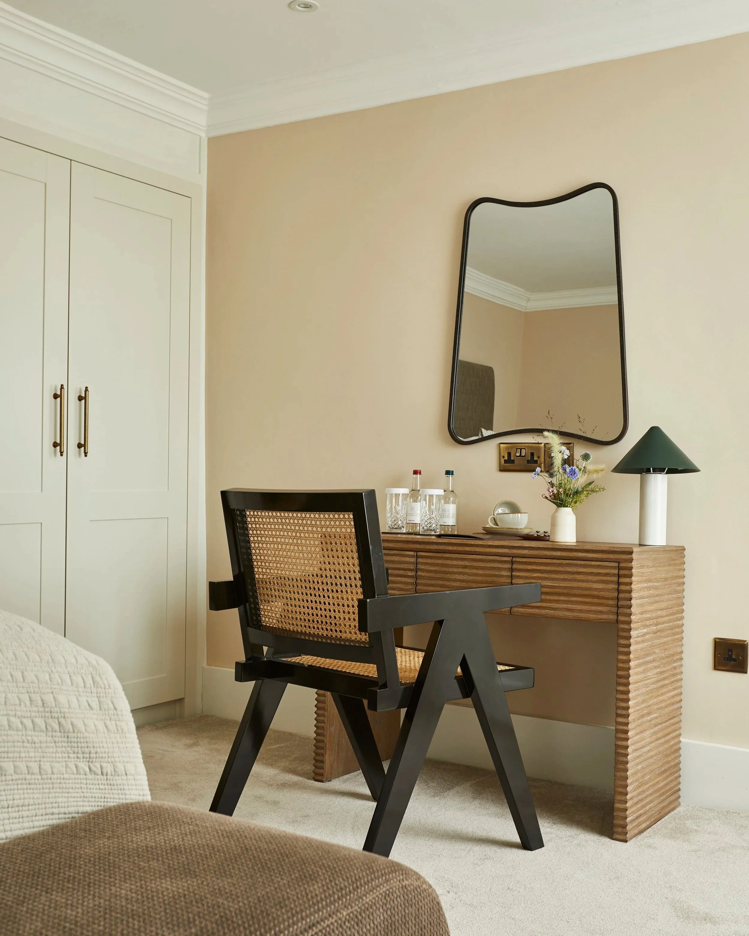 A corner of a bedroom with a wooden sideboard, a black chair with woven rattan backrest, a mirror, a green lamp, and a vase with flowers.