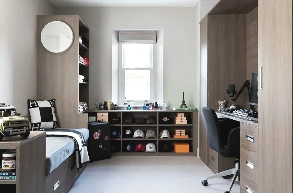 A bedroom with a single bed, desk, and storage units. The bed has a black and white striped blanket, pillows, and a toy car. The wardrobe and shelves are made of light wood in the Home Counties