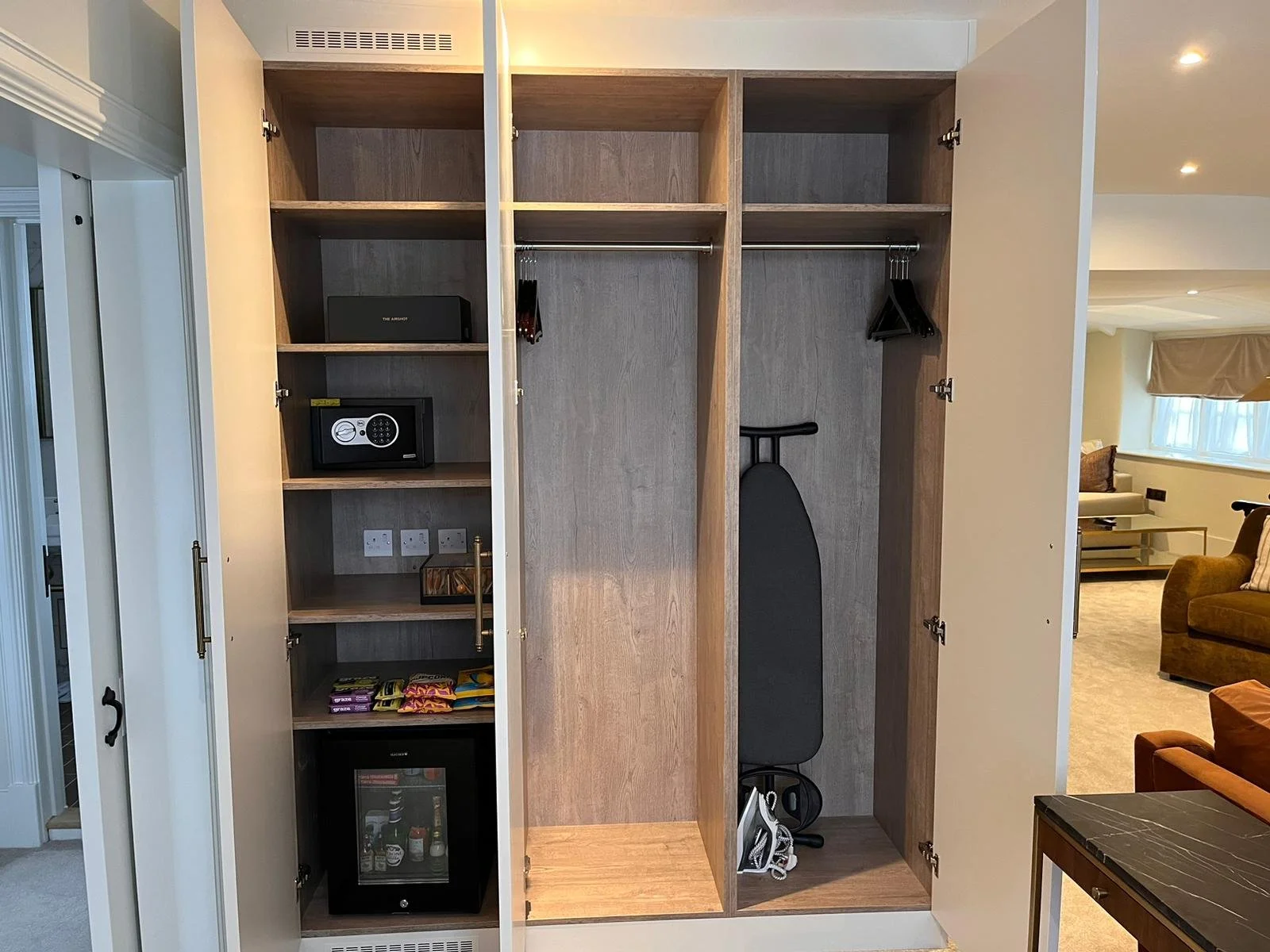 Empty wooden wardrobe with shelves, a safe, an iron, and a mini fridge within a room.