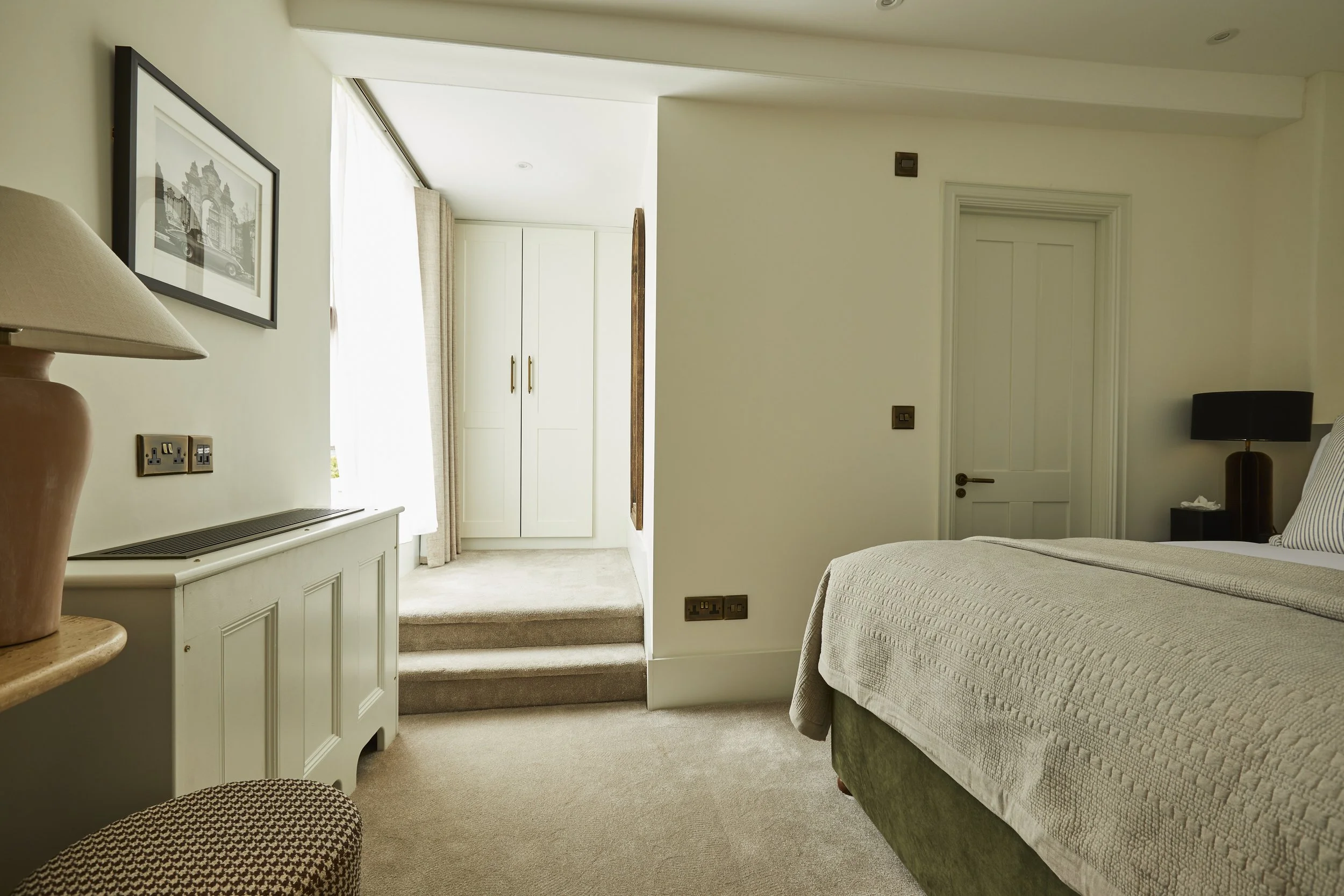 A hotel room with a bed on the right, bedside table with a lamp, a white cabinet on the left, a framed picture on the wall, a window with curtains, and closet doors in the background.