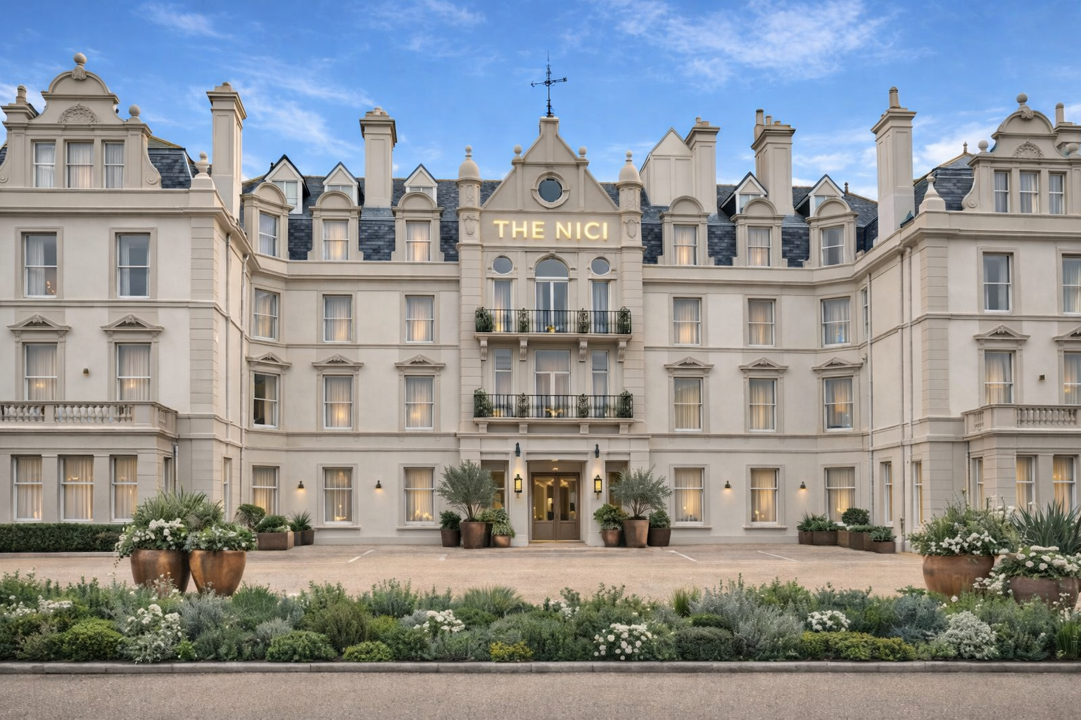 A grand hotel building with a classic European architectural style, beige exterior, multiple windows with white curtains, balconies with railings, and a sign reading 'THE NICI' on the top. The entrance is flanked by potted plants and decorative lighting, with a landscaped garden in the foreground.
