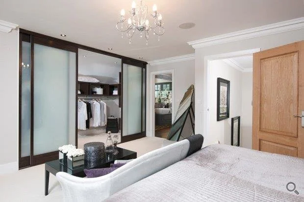 Bedroom with a chandelier, a bed with a padded headboard, a black side table with decorative items, a large mirror, a closet with frosted glass sliding doors, and a doorway leading to another room.