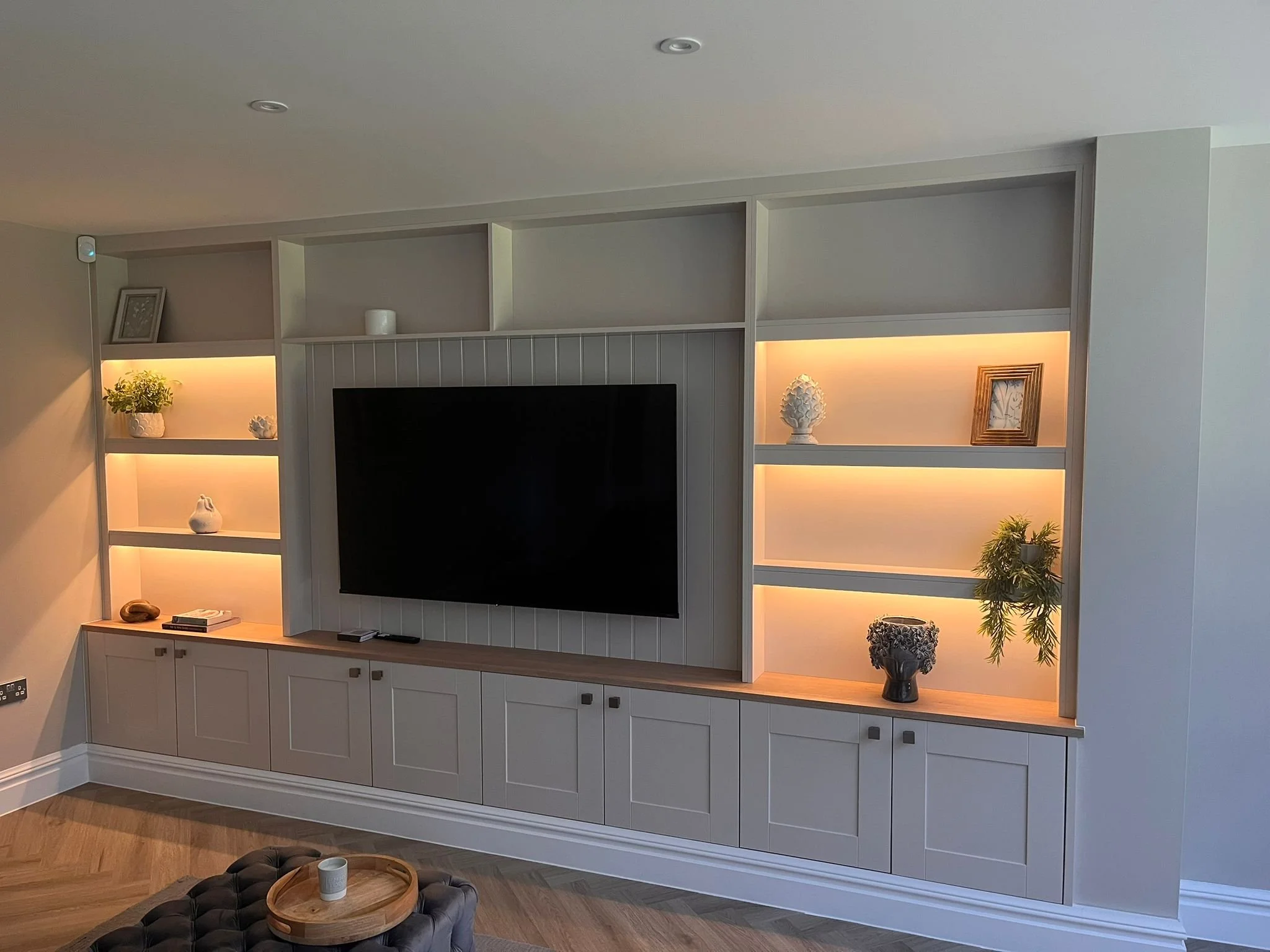 Living room with a wall-mounted flat-screen TV, built-in white shelves with decorative items, and warm LED lighting.