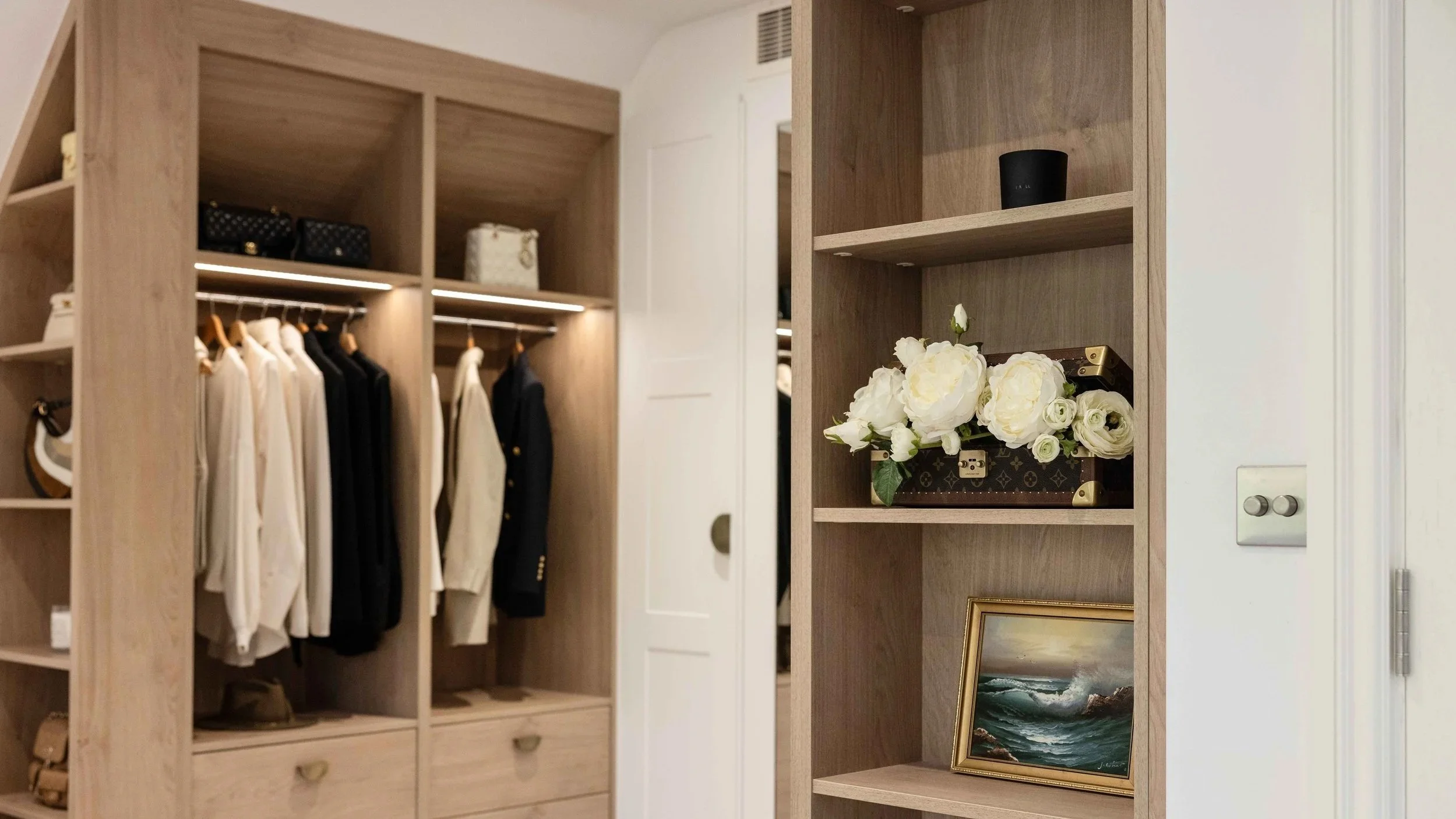 Bedroom closet with clothes hanging on a rod, and shelves with decorative items, a vase of white flowers, and a framed picture of an ocean scene.