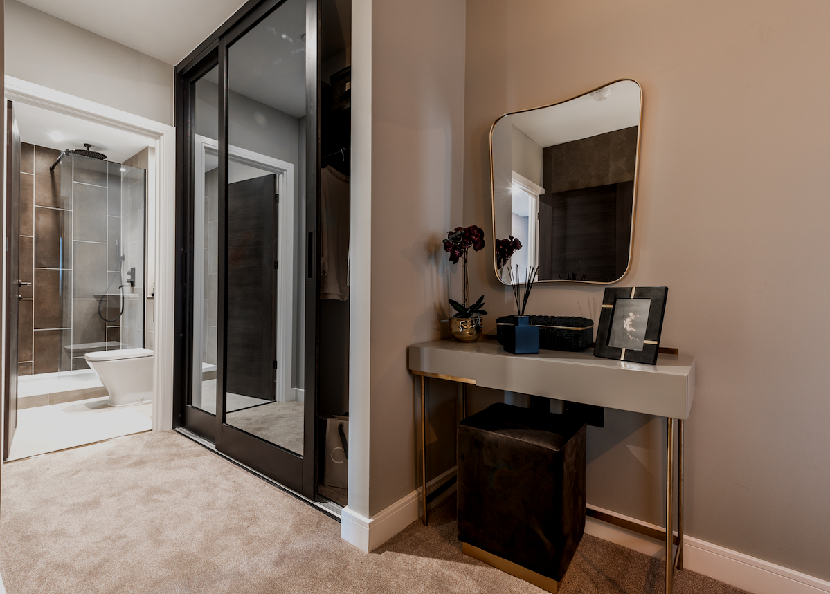 A cozy hotel or apartment bedroom area shown with a small desk, mirror, decorative items, and a nearby bathroom with a glass shower enclosure and toilet.