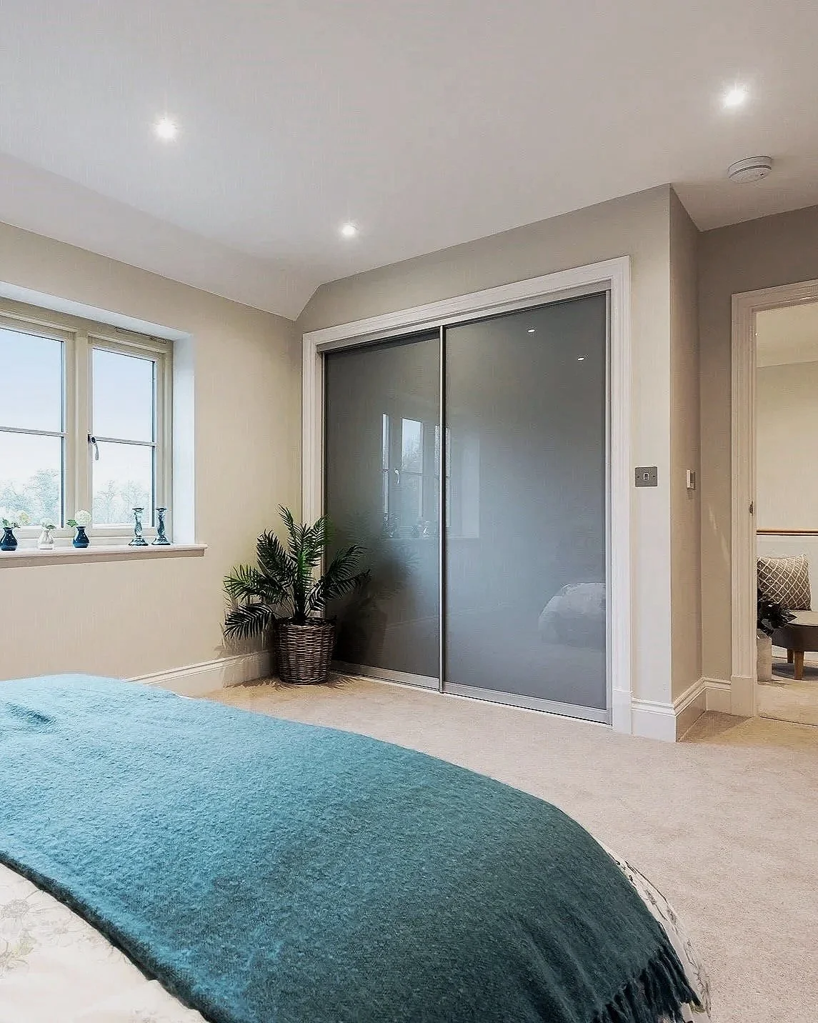 A bedroom with a window, a potted plant, sliding mirrored closet doors, and part of a bed with a teal blanket.
