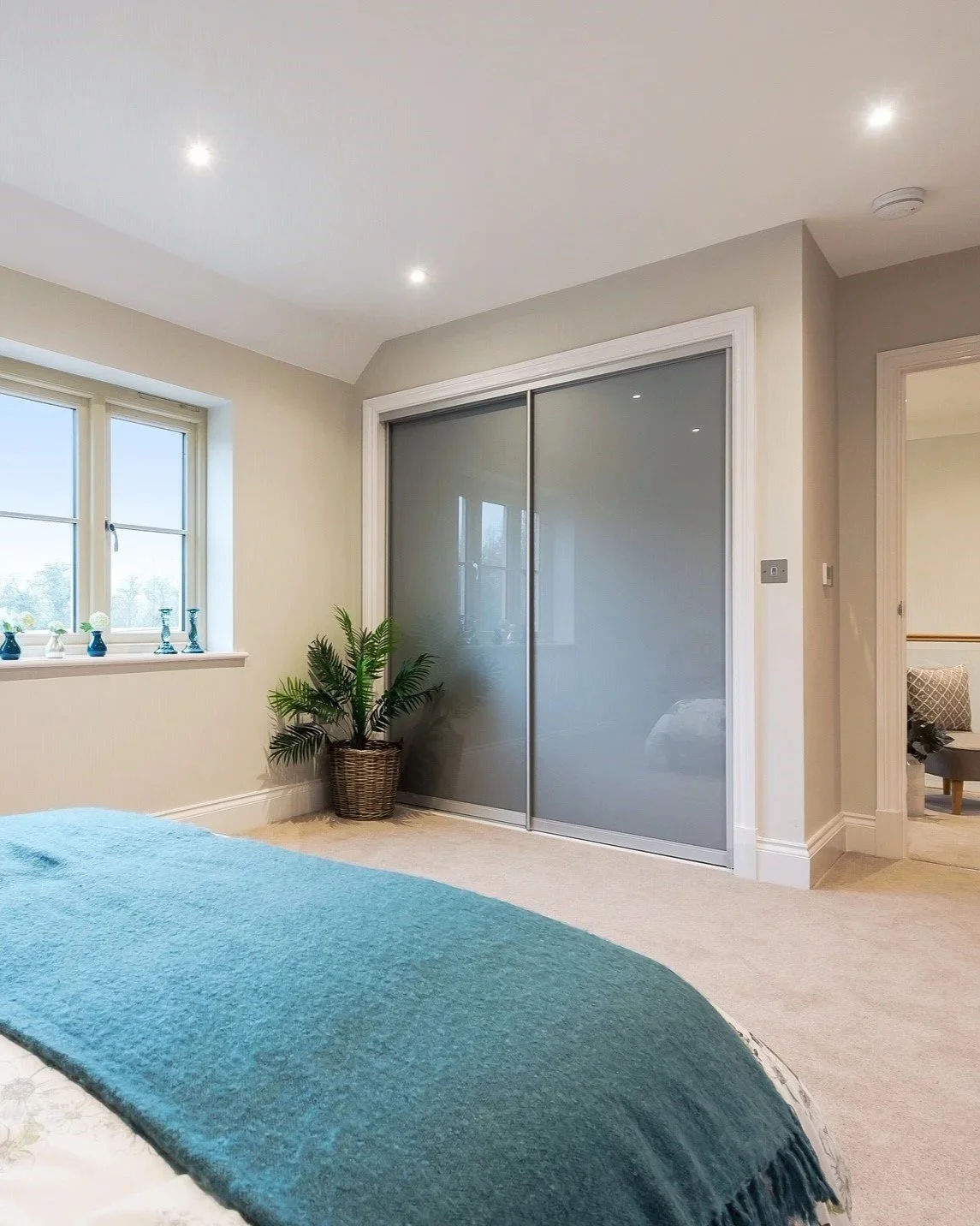 A bedroom with a sliding closet door, a window with decorative vases, a potted plant, and part of a bed with a teal blanket.