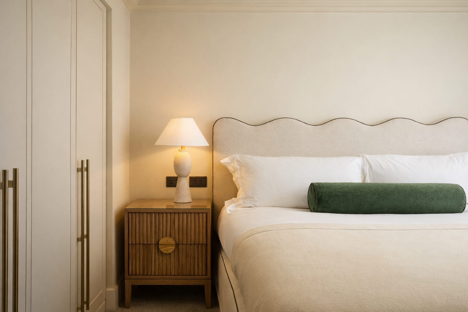 A neatly made bed with white pillows and a green bolster pillow, a beige upholstered headboard, next to a wooden bedside table with a textured white lamp, and a white wardrobe with gold handles in a hotel room.