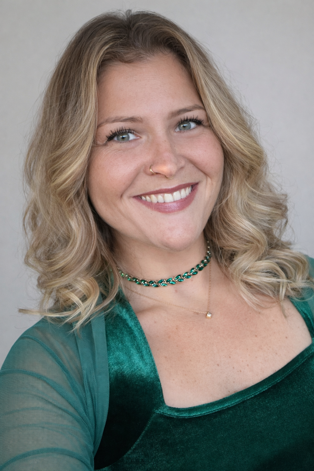 A woman with blond, curly hair smiling, wearing a green velvet top and a green choker, with a small necklace, and a nose ring.