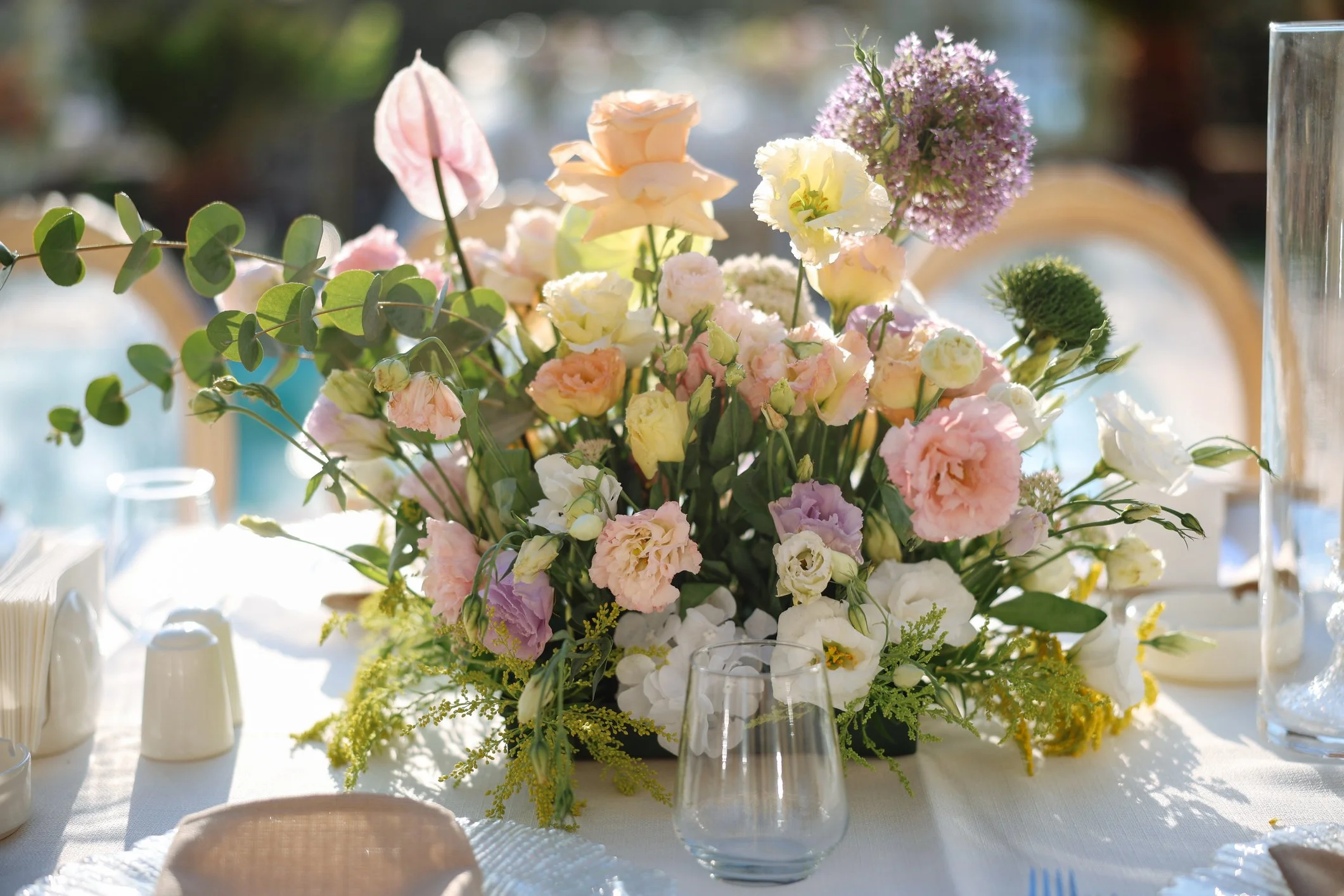 A floral centerpiece with pink, white, and purple flowers on a white tablecloth at a sunny event.